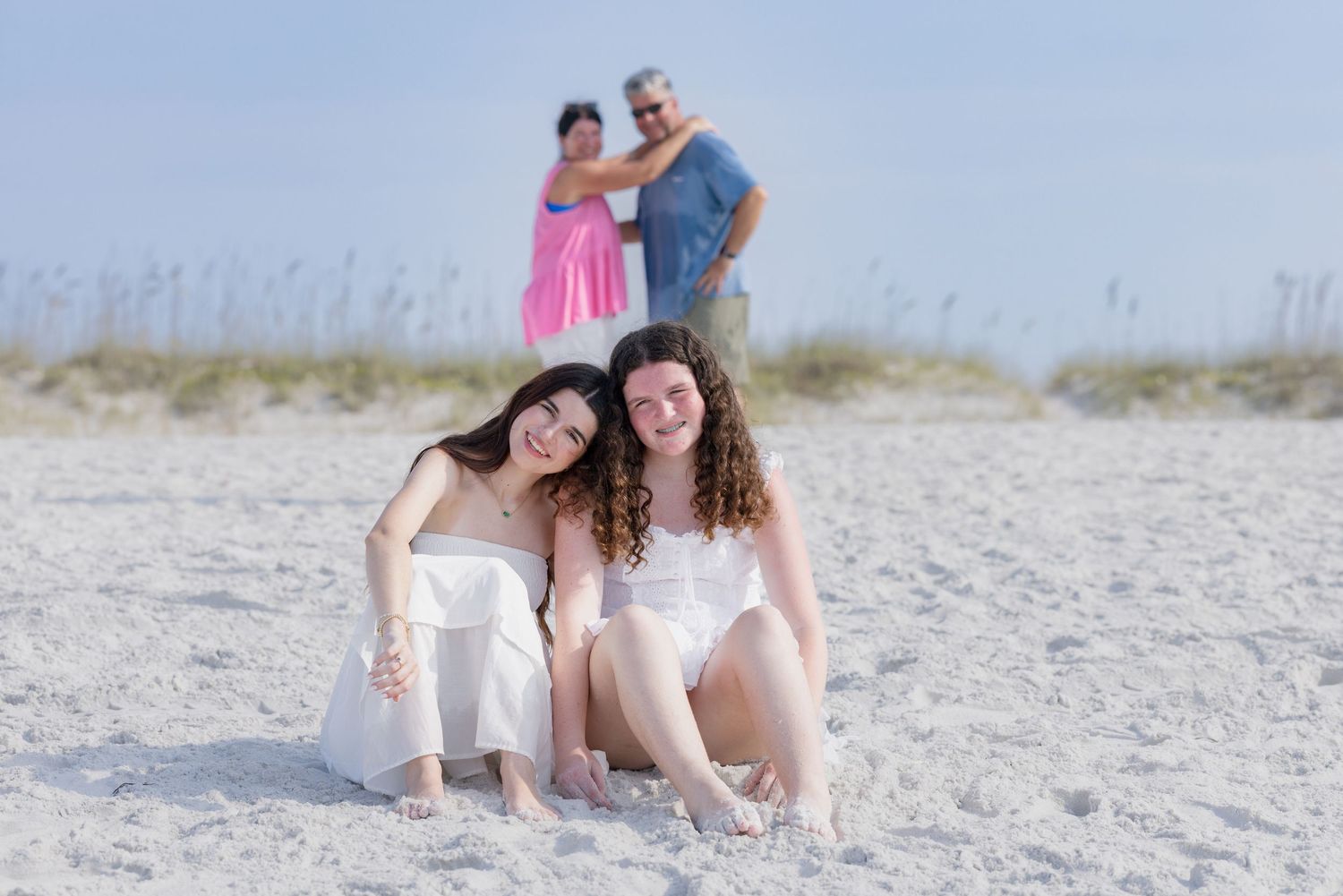 Two young women sit on a white sandy beach smiling, with a couple standing in the background near sand dunes.