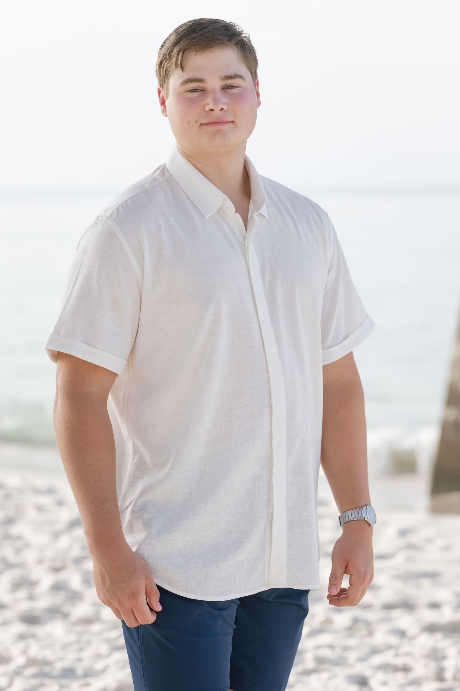 Young man in white button-up shirt and navy pants standing on beach with ocean in background.