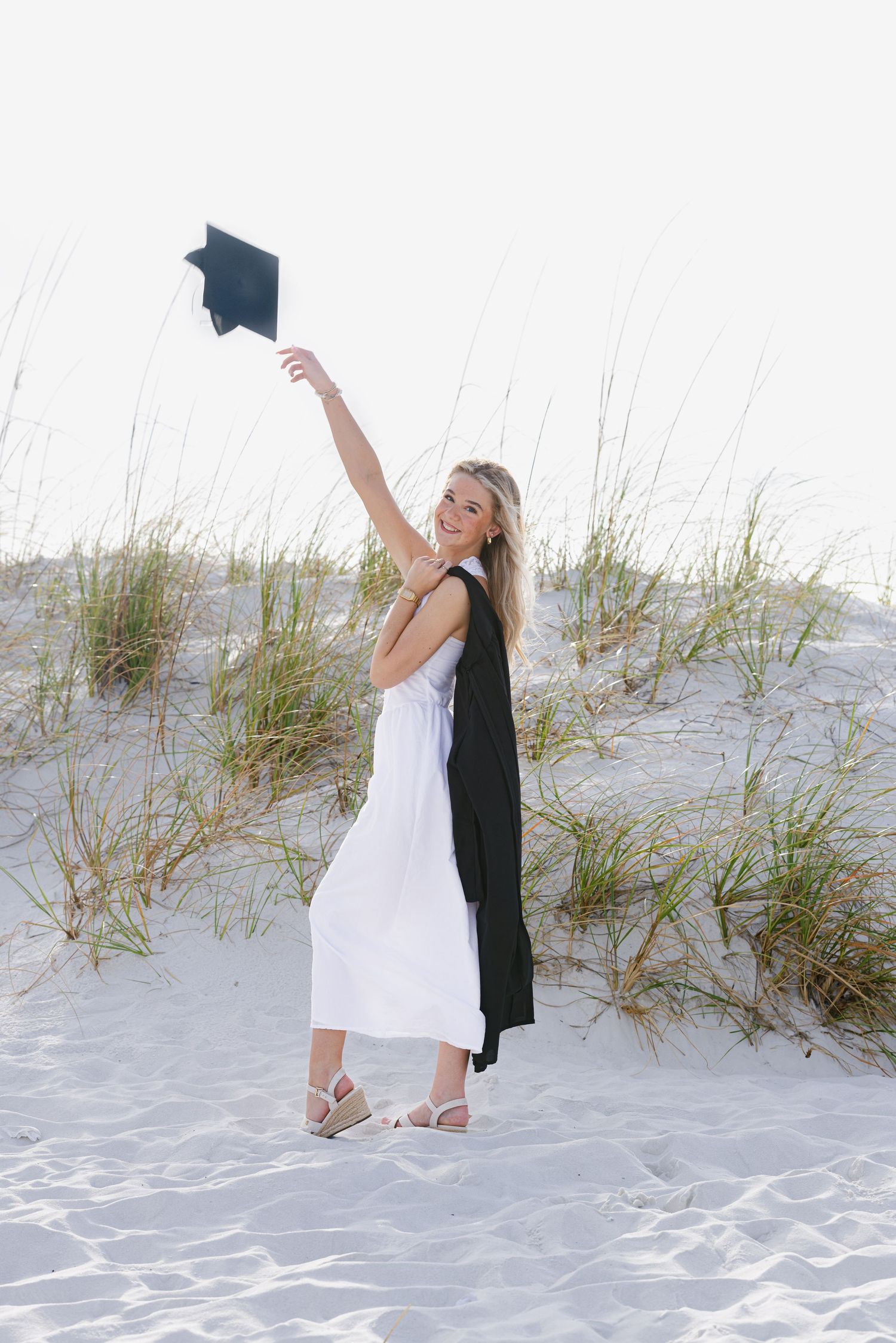 Smiling blonde woman in white dress and black graduation gown raises cap on sandy beach dunes.
