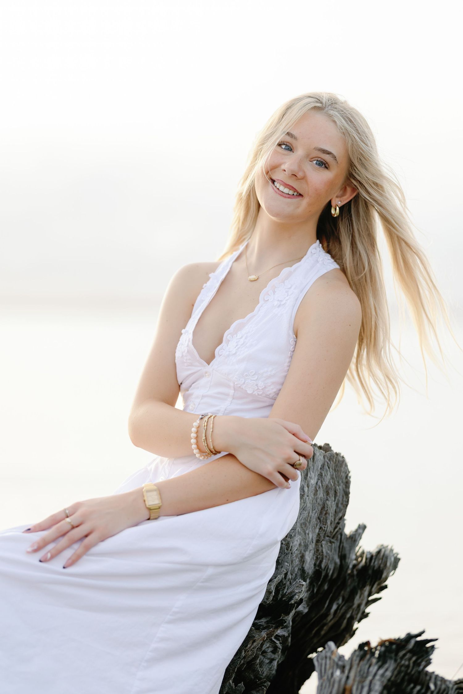 Smiling blonde woman in white lace dress sitting on driftwood at a beach, wearing gold jewelry.