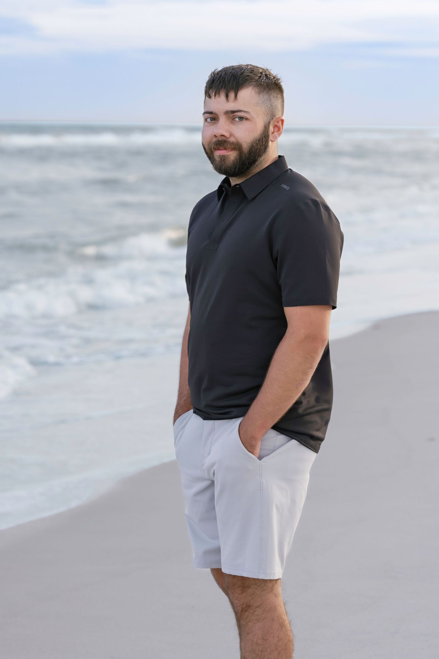 Young bearded man in black polo shirt and white shorts standing on sandy beach with ocean waves behind him.