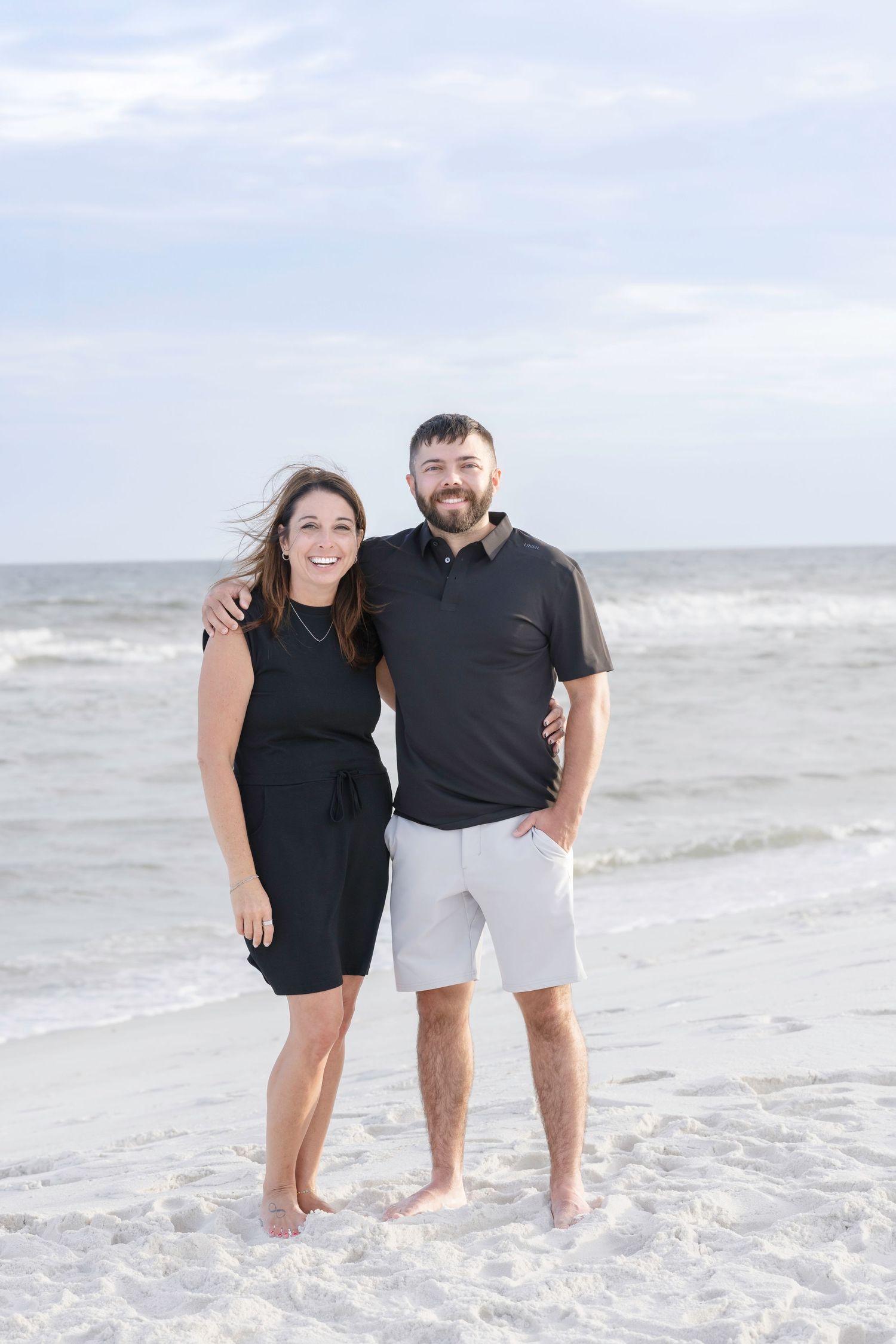 Couple smiling together on a sandy beach, woman in black dress and man in black shirt and gray shorts.