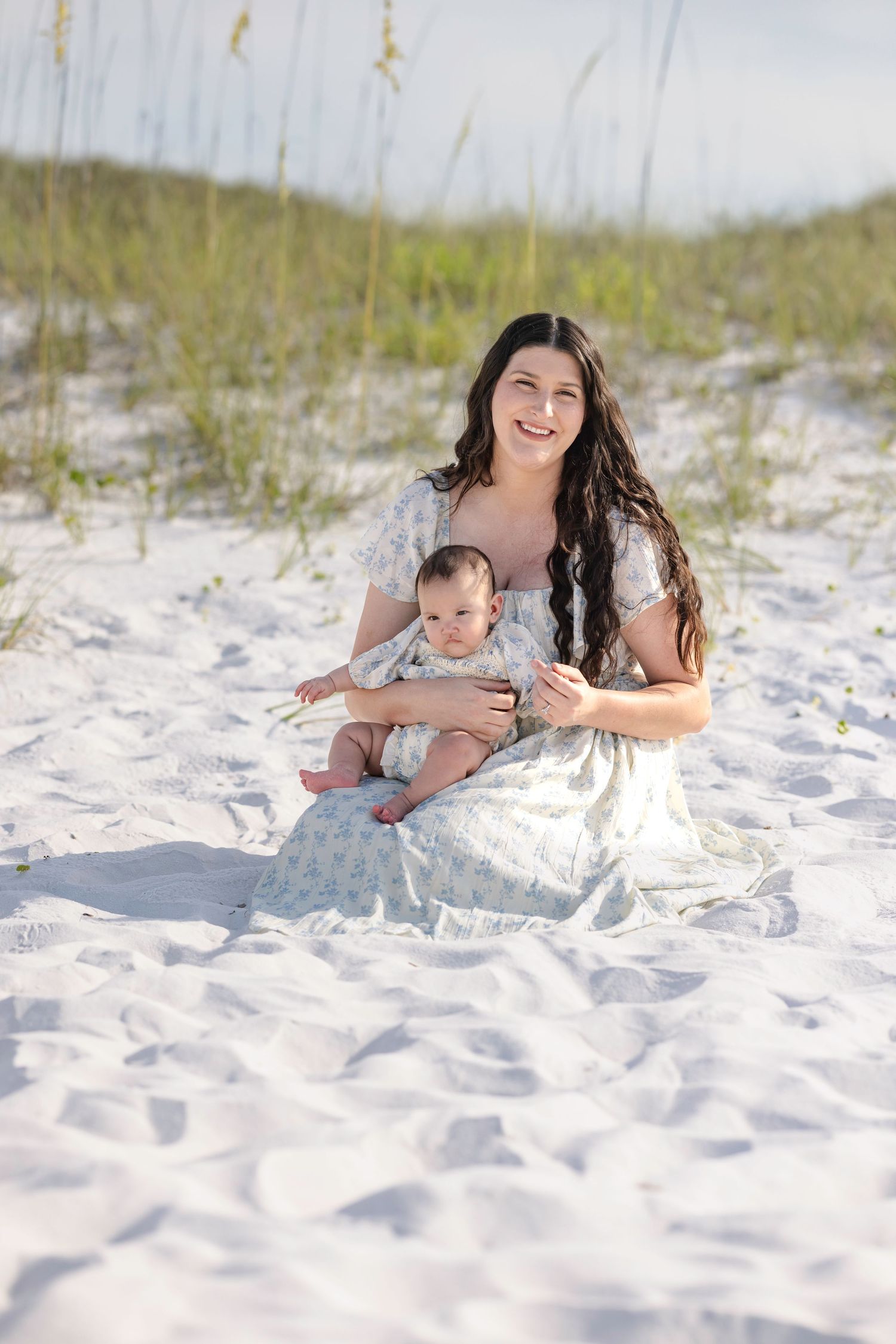 A smiling mother in a floral dress sits on white sand dunes holding her baby, with sea oats in the background.
