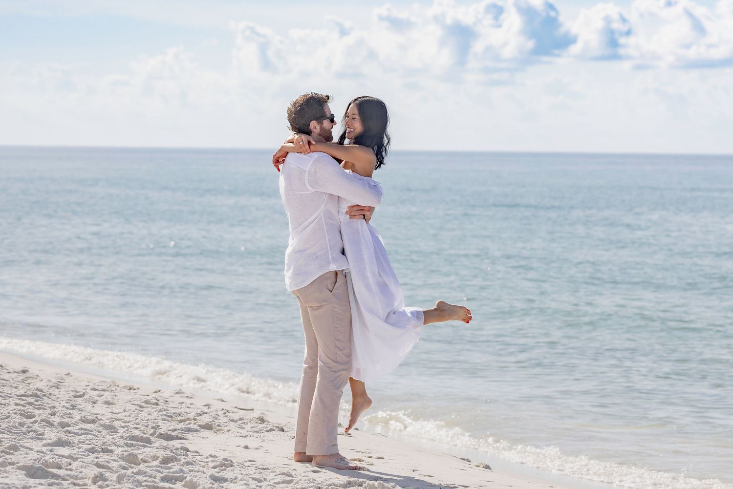 Romantic couple embracing on a white sand beach, man lifting woman as they share a joyful moment by the sea.