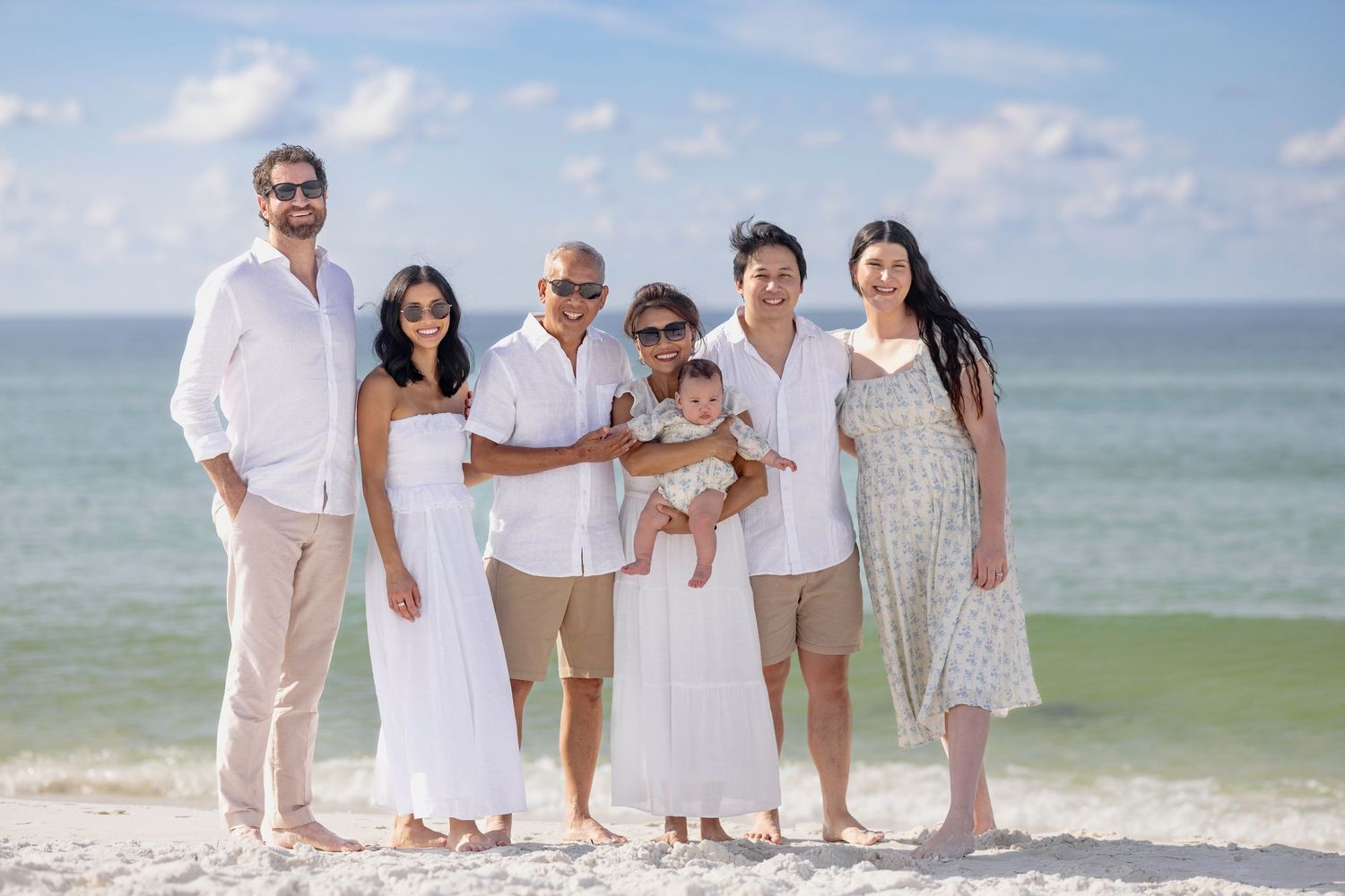 Multi-generational family posing together on a sunny beach, dressed in white outfits with ocean waves in the background.