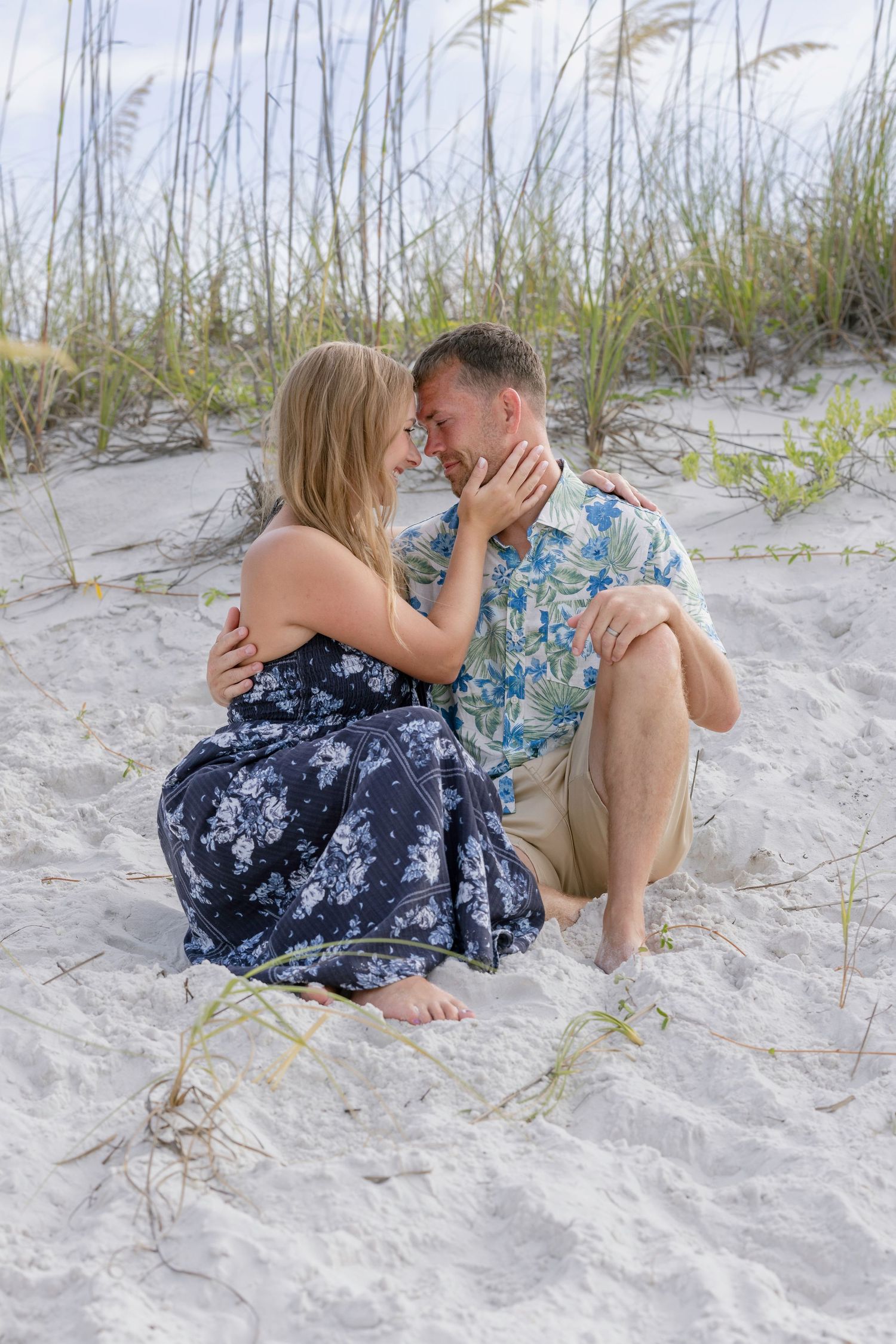 Couple sitting on white sand dunes, sharing a romantic moment near sea oats at the beach.