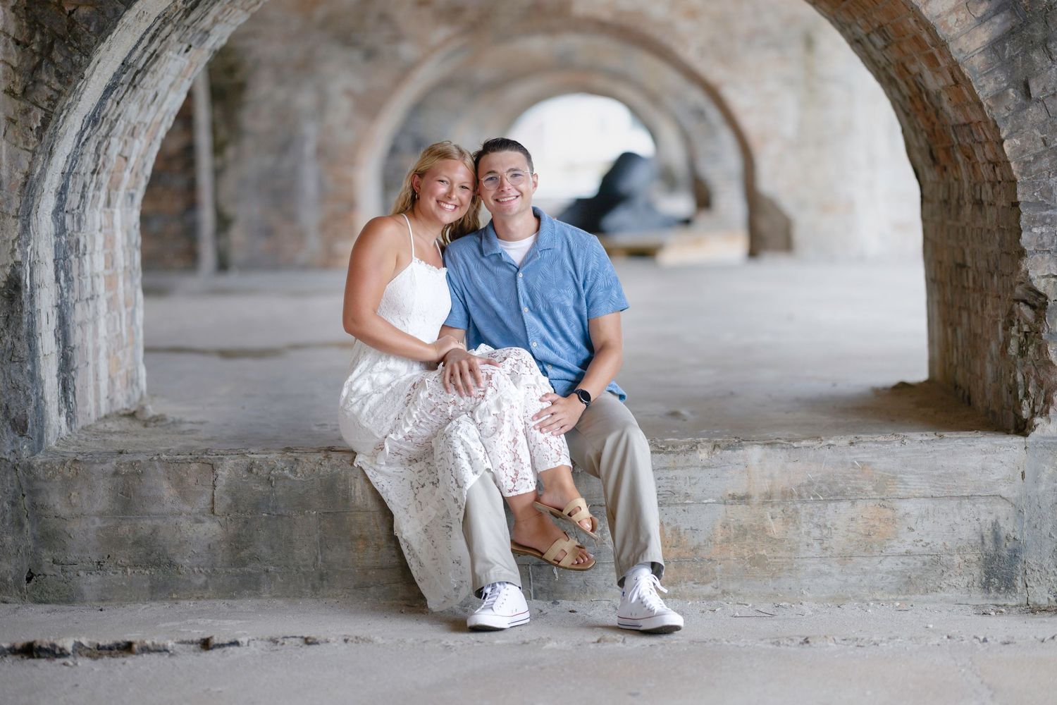 Couple sitting together under stone arches, woman in white dress and man in blue polo shirt, smiling warmly.
