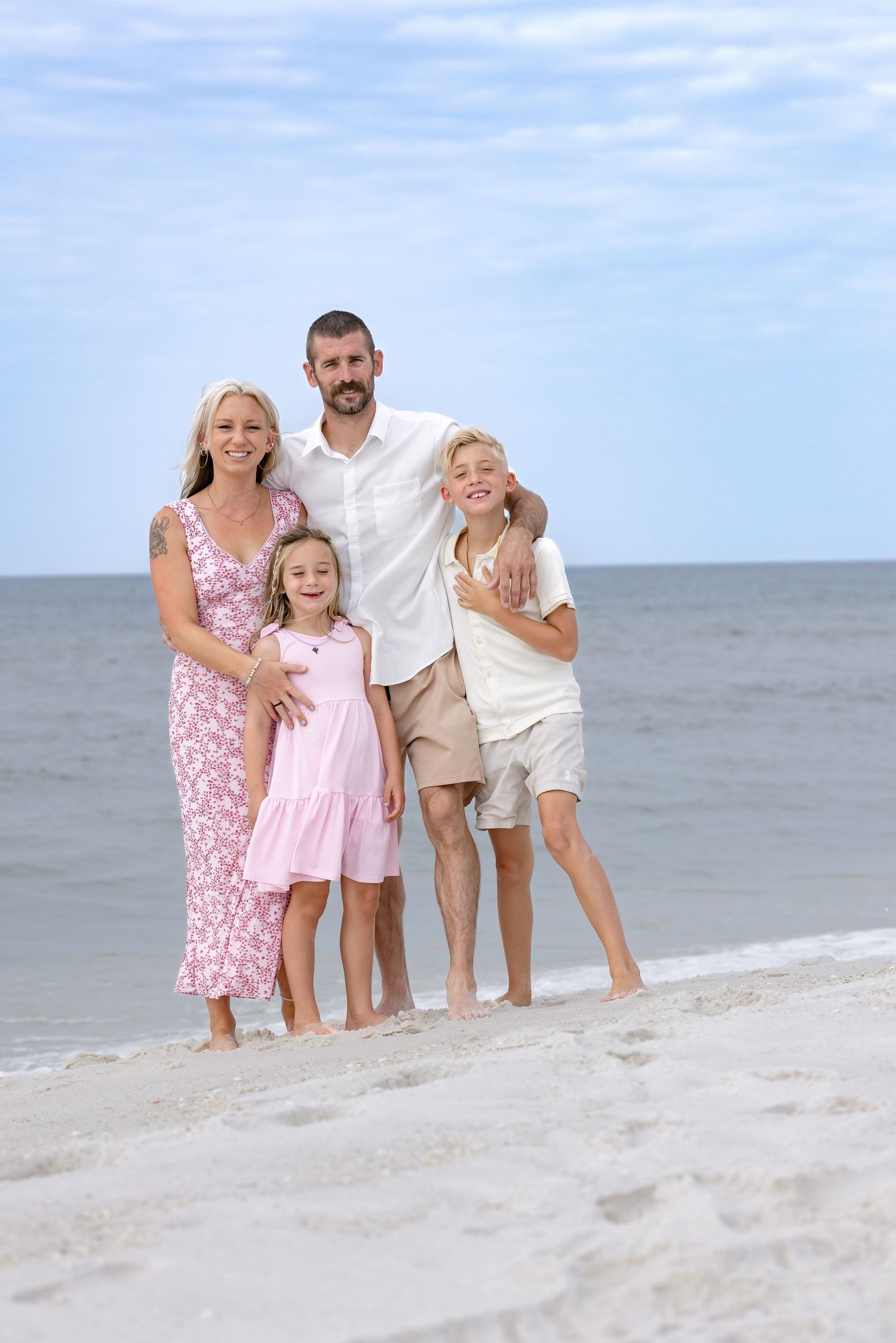 Happy family of four posing together on a sandy beach with calm ocean waves in the background on a sunny day.