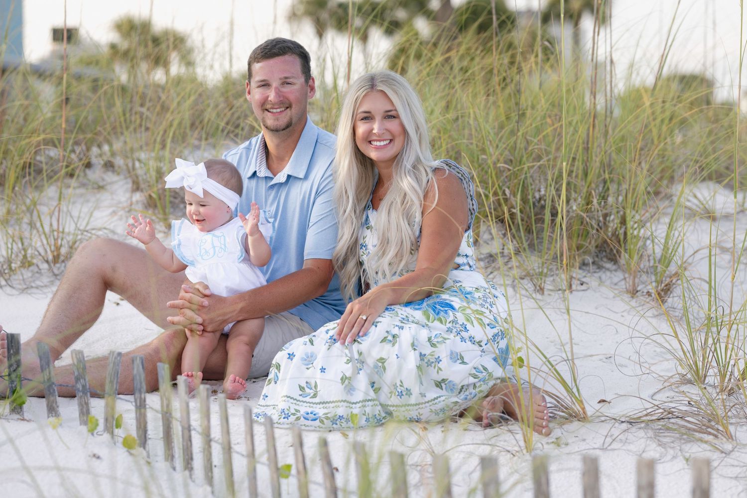 Young family with baby girl sitting on white sand beach dunes, smiling during a sunny beach photo session.