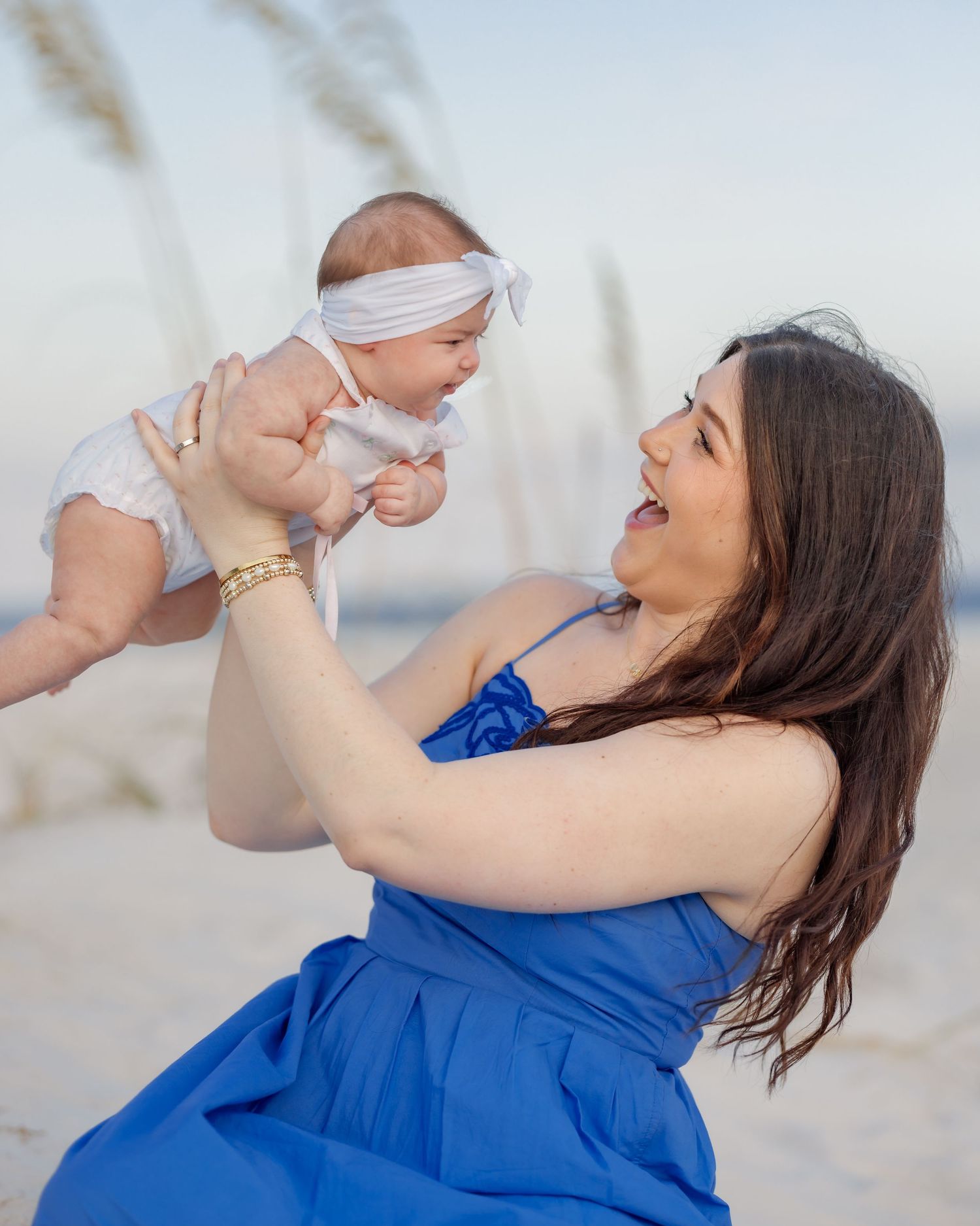 Joyful mother in blue dress lifting laughing baby in white on sunny beach.