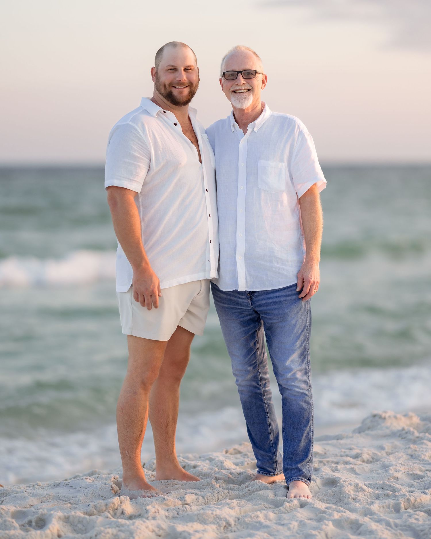Two smiling men in white shirts pose together on a sunny beach.