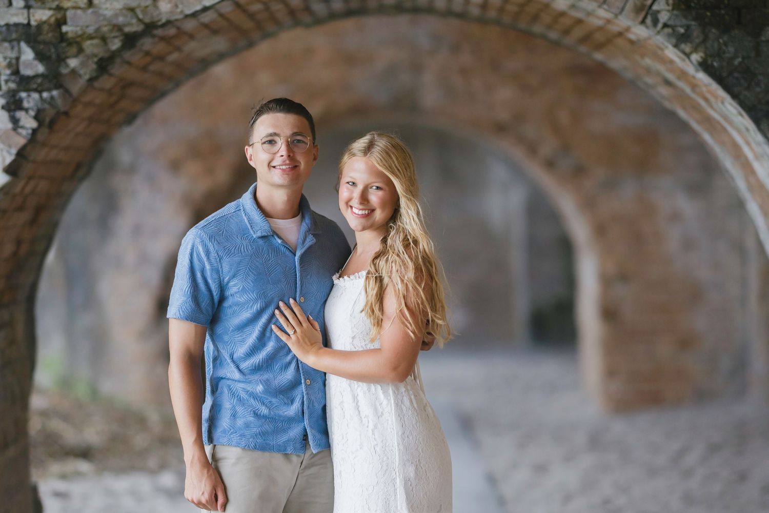 Couple smiling under stone arches, man in blue shirt and woman in white dress, romantic outdoor portrait.