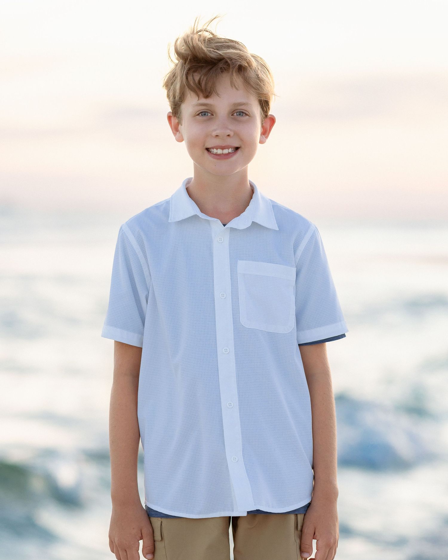 Boy smiling at beach wearing white short-sleeve button-up shirt with chest pocket and khaki shorts.