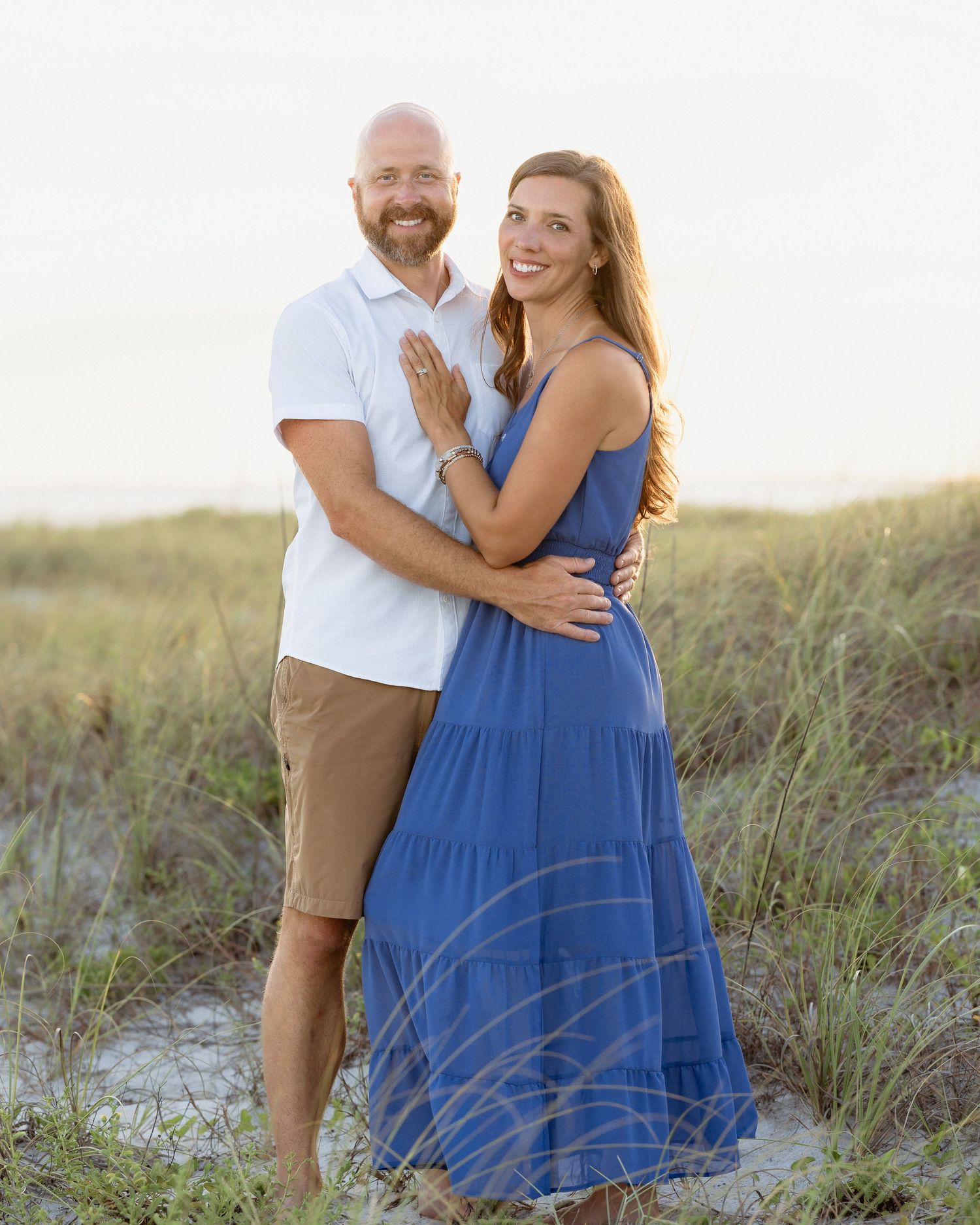 Couple embracing on sandy beach dunes at sunset, woman in blue maxi dress, man in white shirt and khaki shorts.