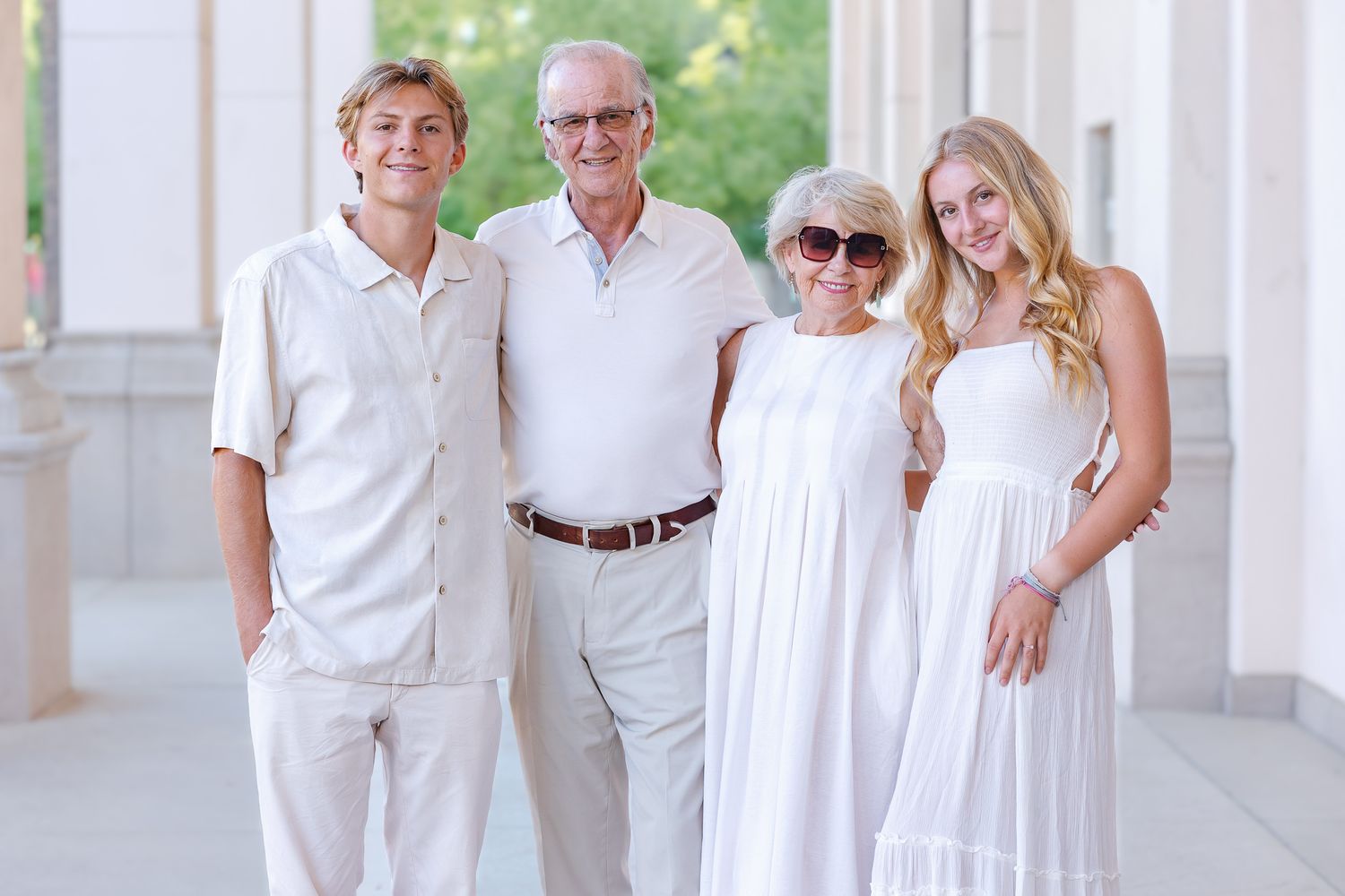Family of four posing together in white outfits, smiling outdoors near elegant columns.