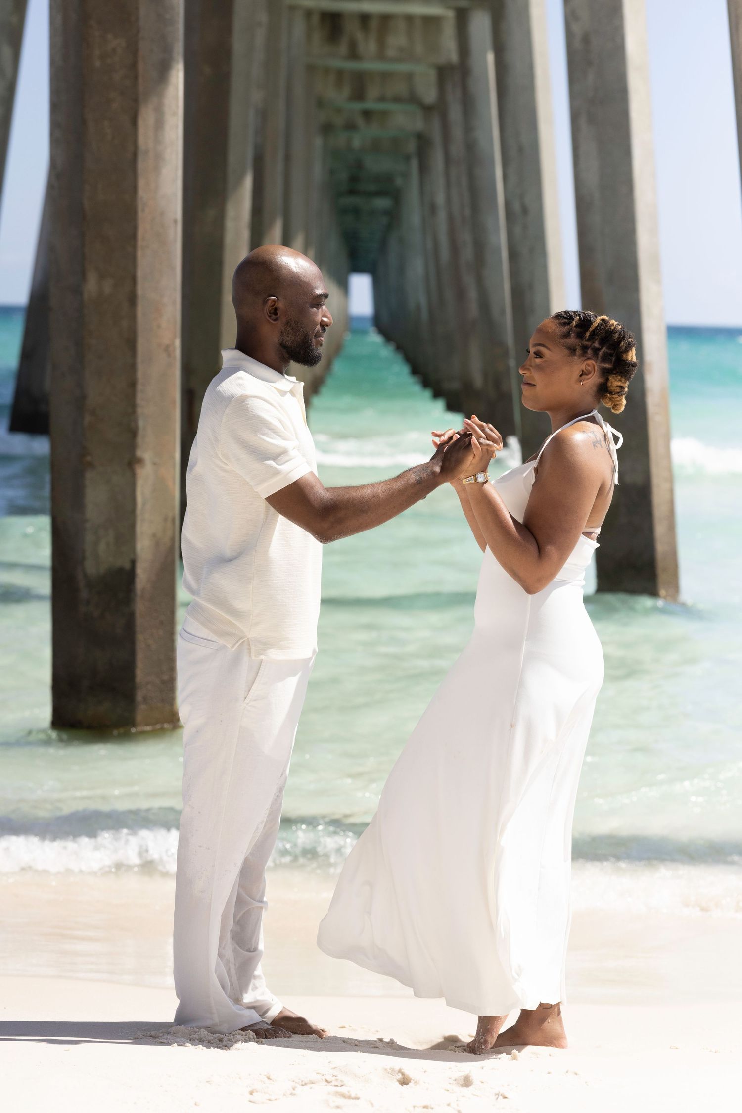 A couple in white attire holds hands on a sunny beach near a pier with turquoise ocean waves in the background.