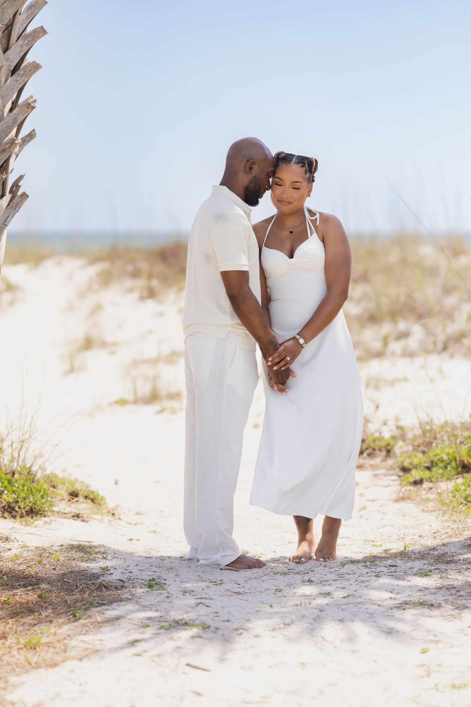 A couple in white outfits holds hands on a sandy beach, sharing an intimate moment near a palm tree.