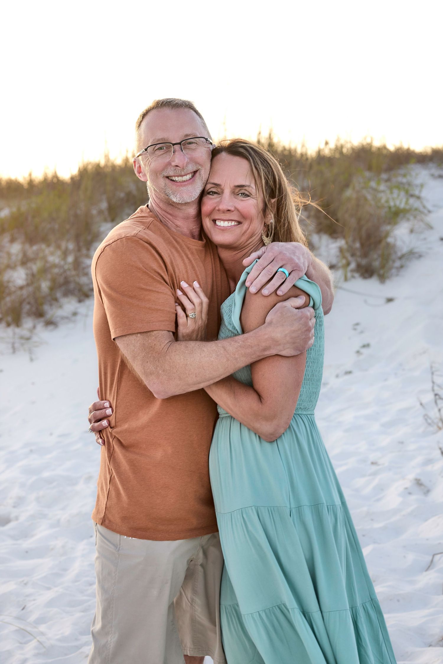 A smiling couple embraces on a white sand beach at sunset, woman in teal dress, man in orange shirt and khaki shorts.