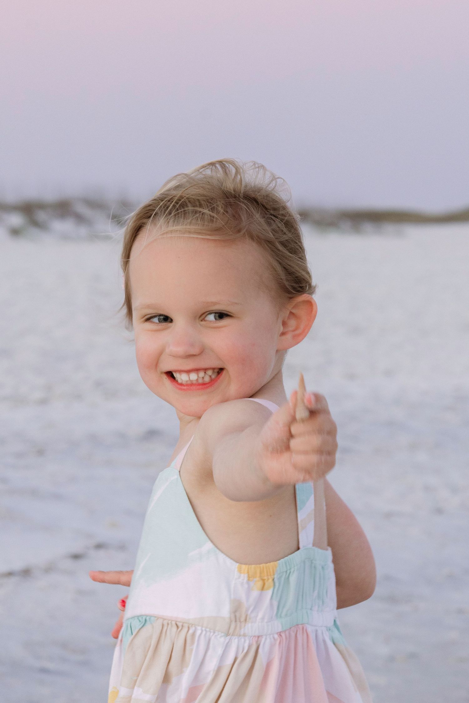 Smiling toddler girl in colorful sundress reaches toward camera on a white sand beach at dusk.