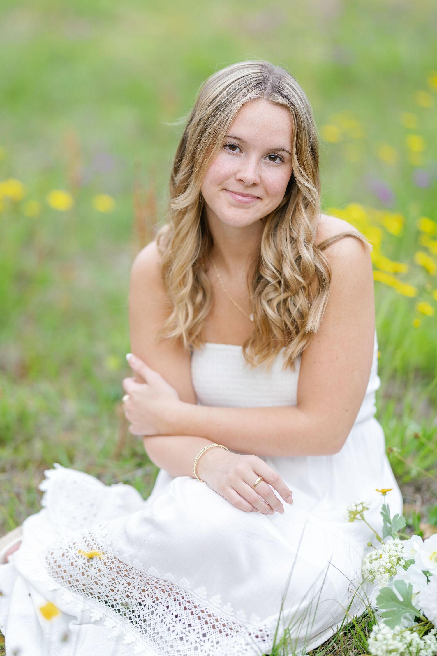 Young blonde woman in white dress sitting in a wildflower meadow, smiling warmly at the camera.
