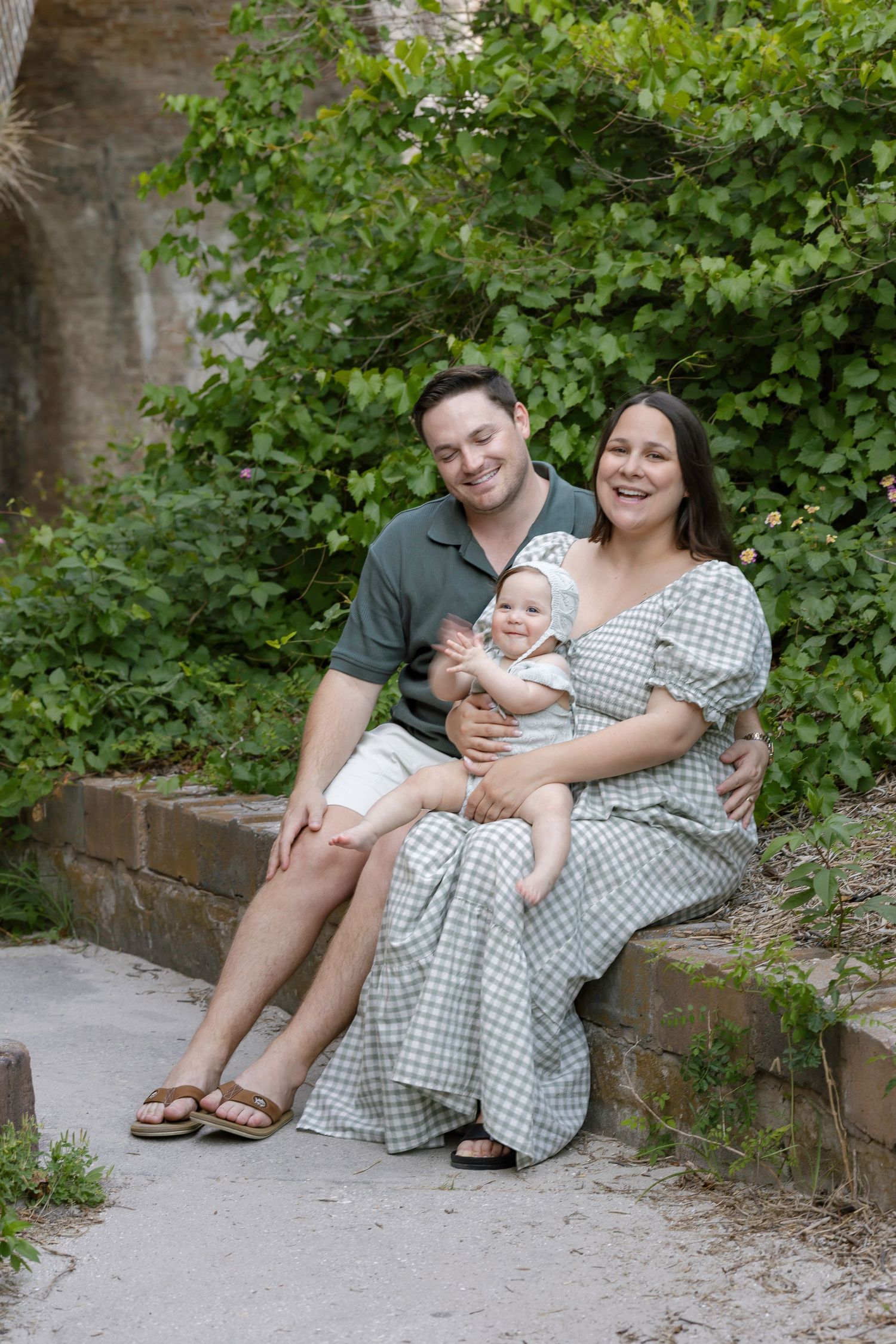 Happy family of three sitting outdoors on stone steps, parents smiling at baby in green leafy garden setting.