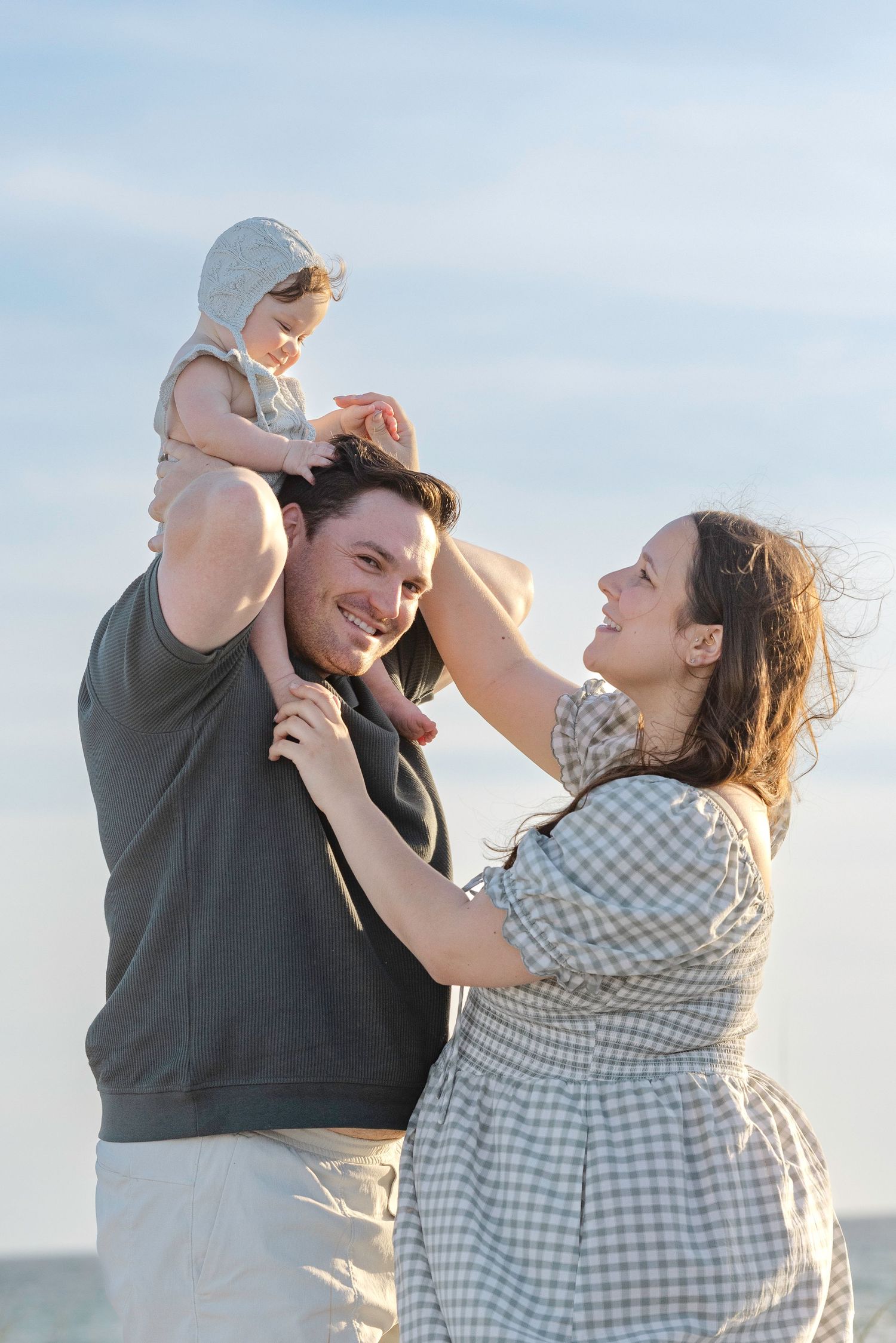 Happy family with father holding baby on shoulders while mother smiles outdoors under blue sky.