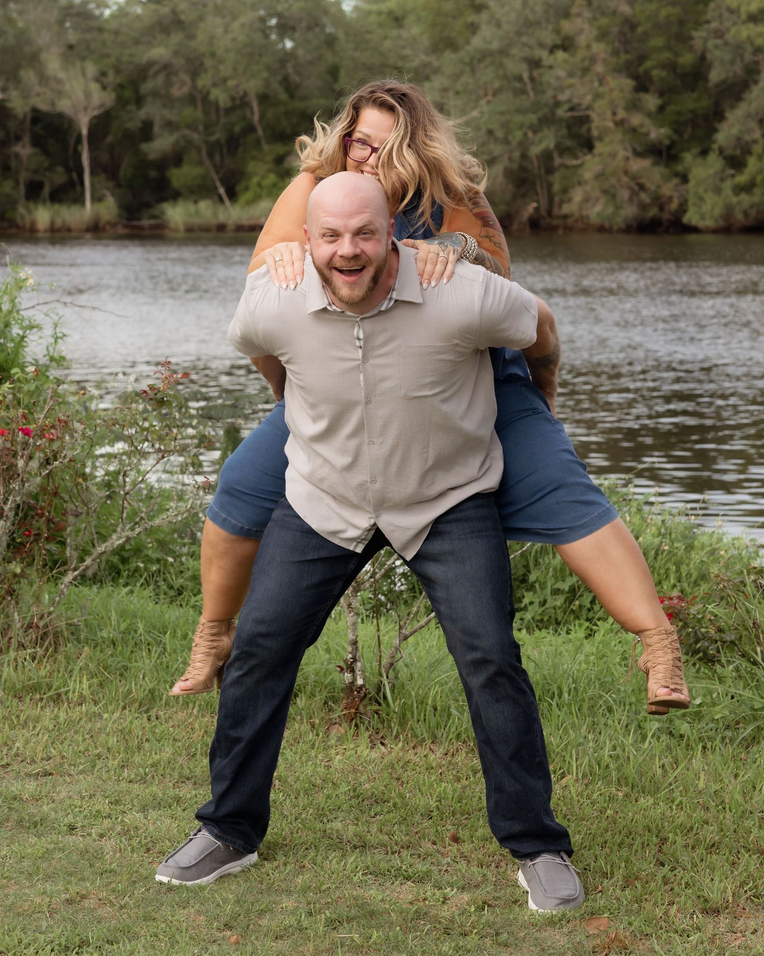 A bald man gives a laughing blonde woman a piggyback ride near a scenic river outdoors.
