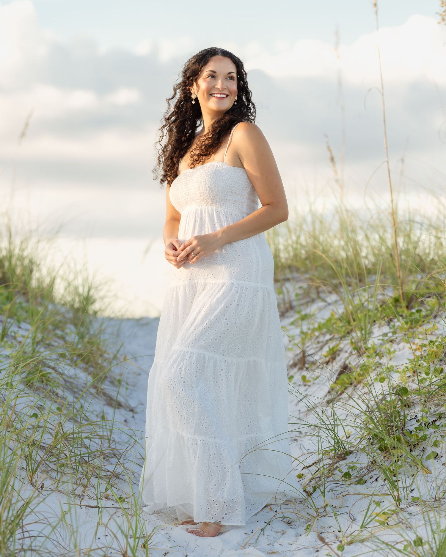 A smiling woman in a white lace maxi dress stands barefoot on a sandy beach with sea oats in the background.