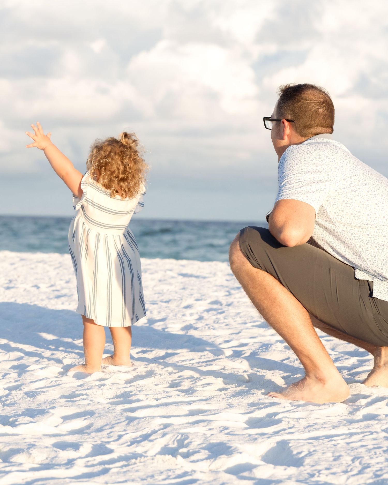 A father crouches beside his curly-haired toddler girl in a striped dress on a sunny white sand beach.