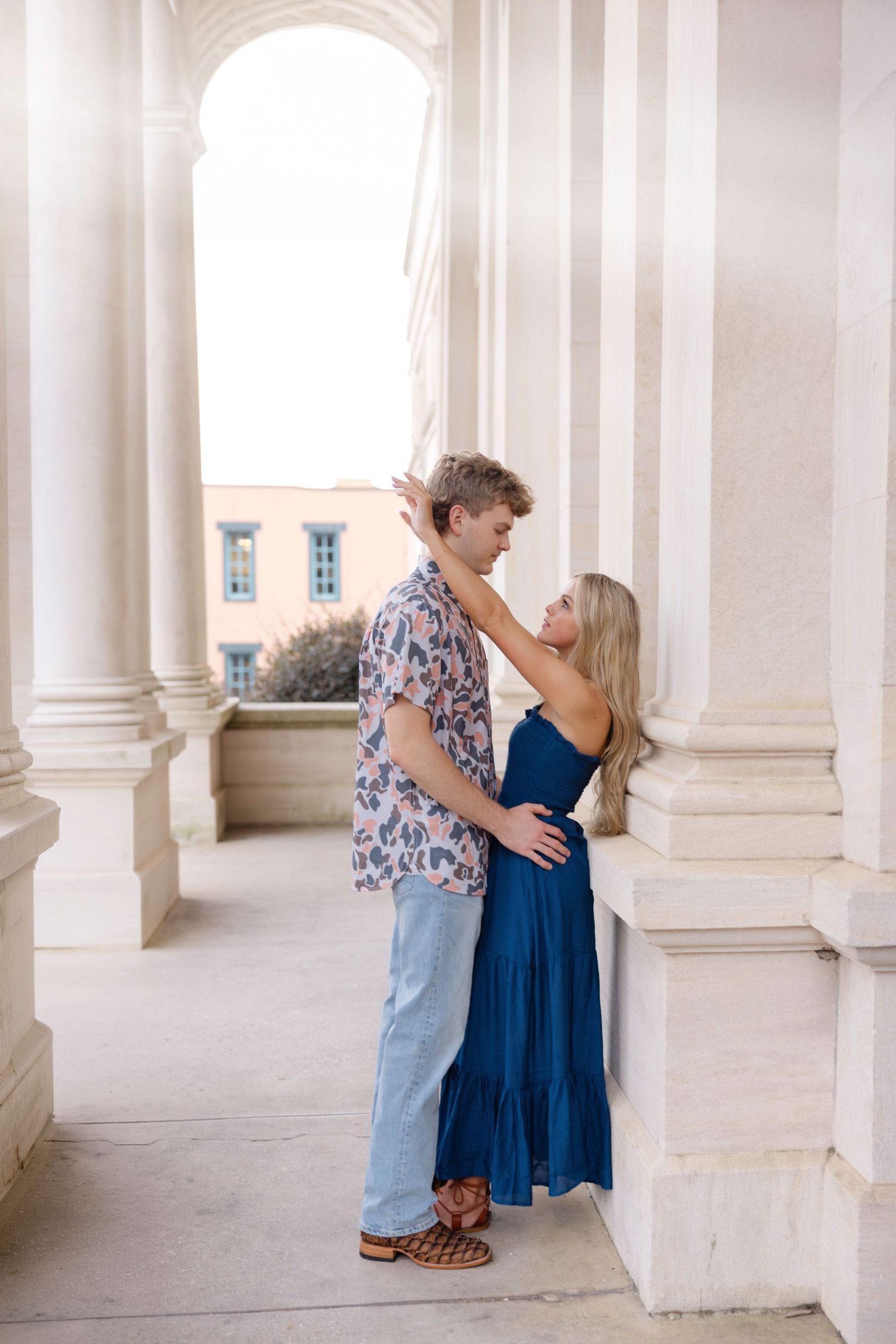 A couple shares an intimate moment in a bright colonnade with white columns and natural lighting.
