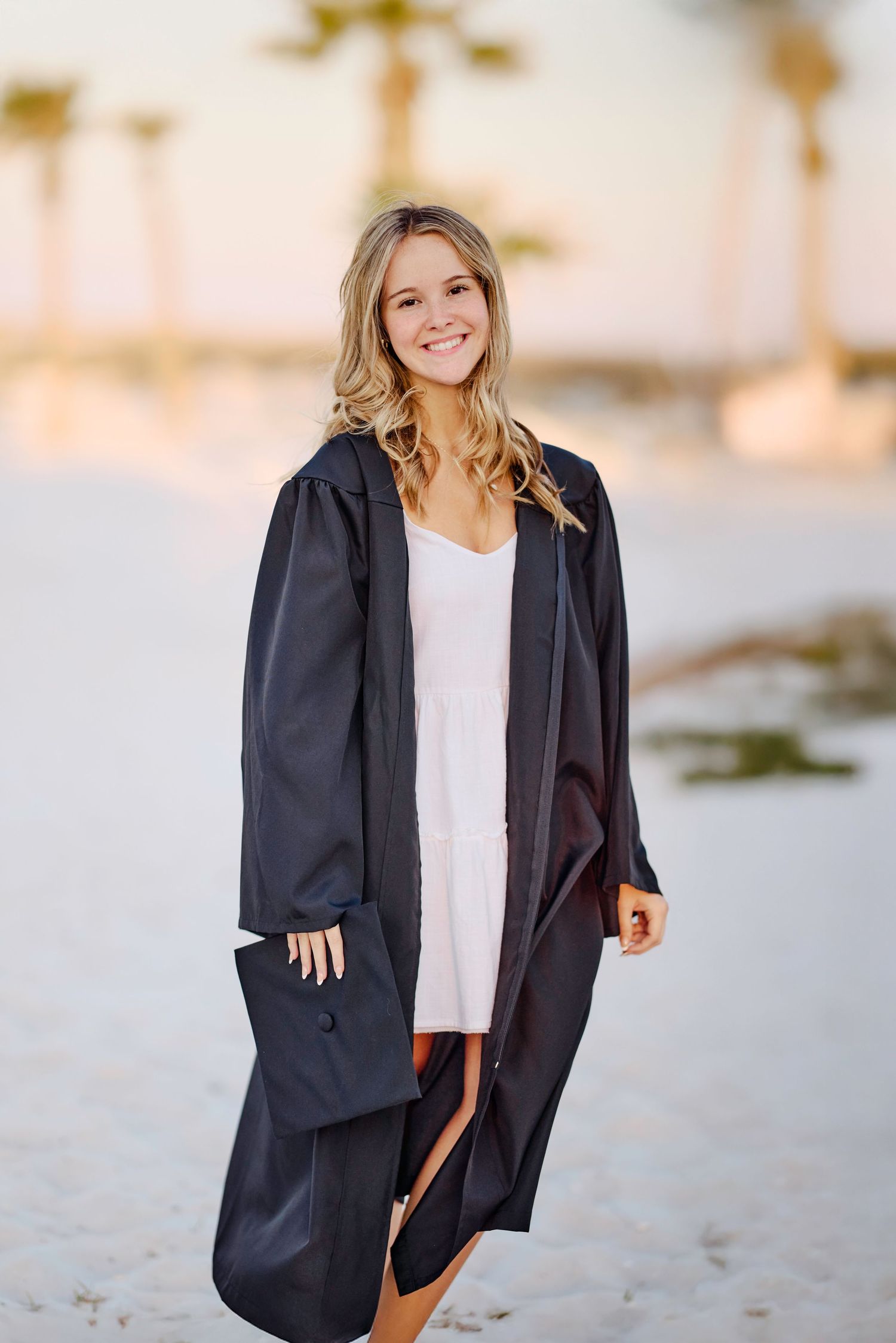 Person wearing a black graduation gown stands smiling on a beach with palm trees in the background at sunset.
