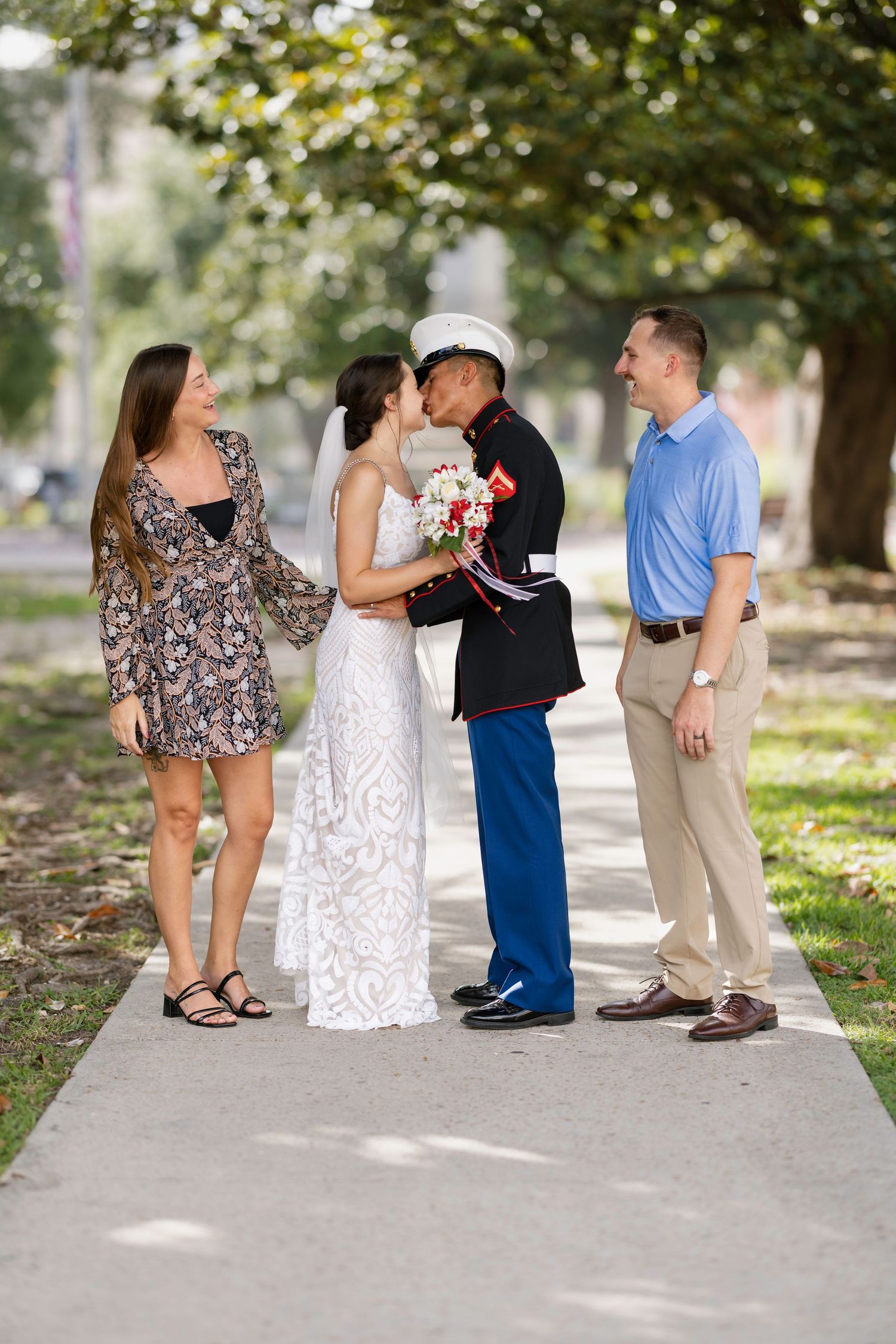 Marine groom kissing bride while two guests smile and watch during outdoor wedding photo session.