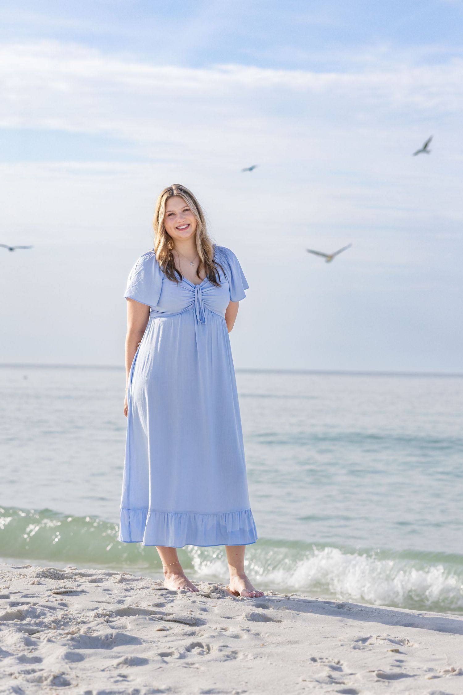 Smiling woman in light blue maxi dress standing barefoot on sandy beach with seagulls flying overhead.