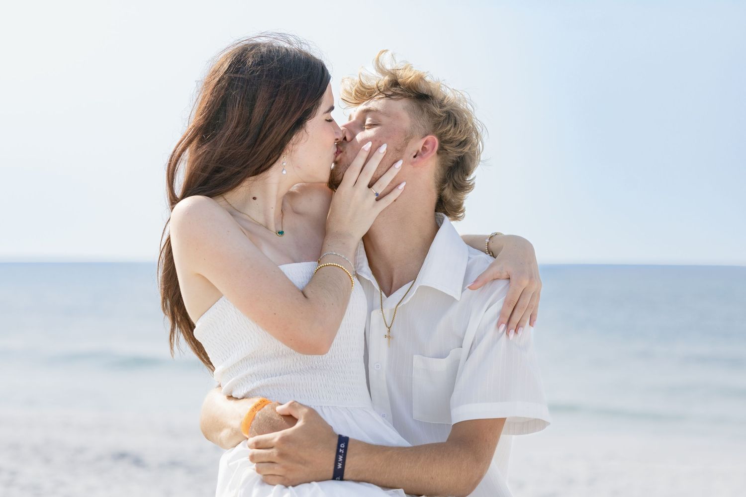 Romantic couple sharing a kiss on a sunny beach, embracing in white outfits with the ocean in the background.