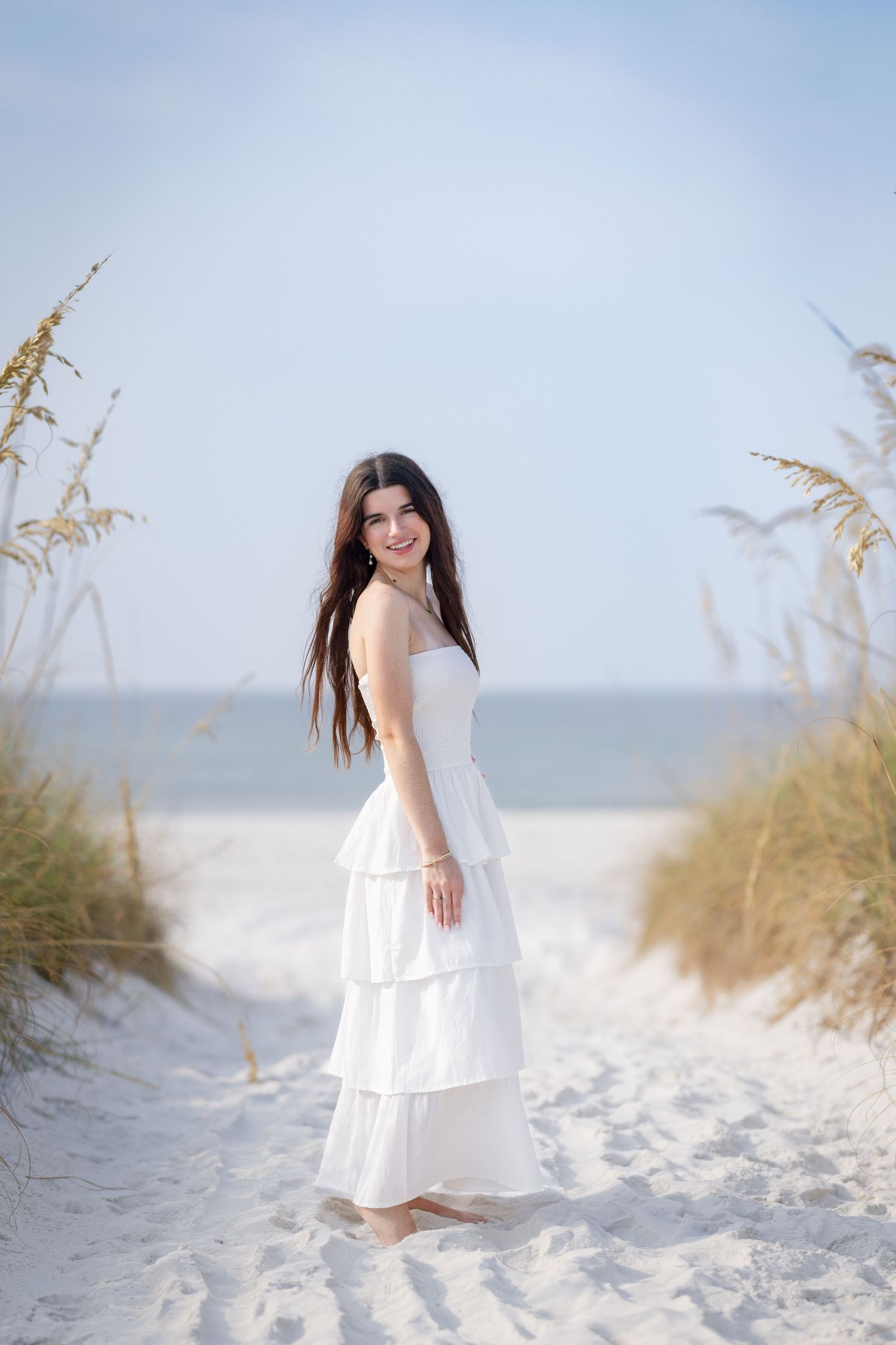 Young woman in white tiered maxi dress standing on sandy beach path framed by sea oats under blue sky.