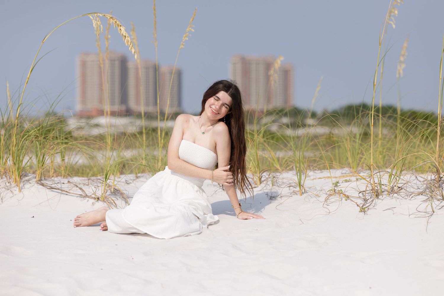 Young woman in white strapless dress sitting gracefully on white sand dunes with city skyline behind.