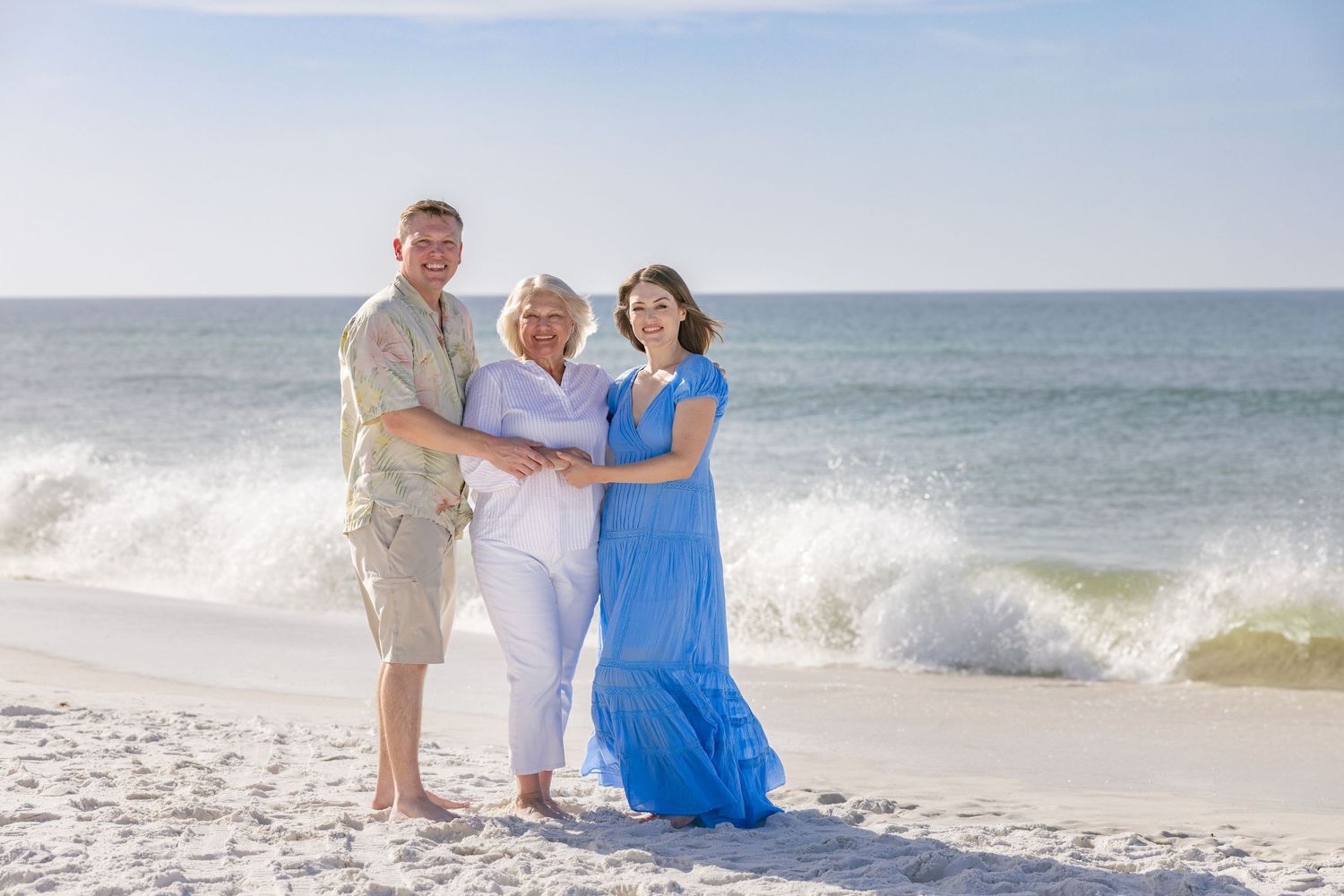 Three people smile together on a sunny beach with ocean waves, one woman in a blue maxi dress.