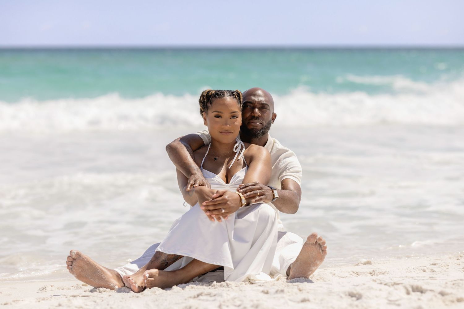 Couple sitting together on a sandy beach, embracing with ocean waves in the background on a sunny day.