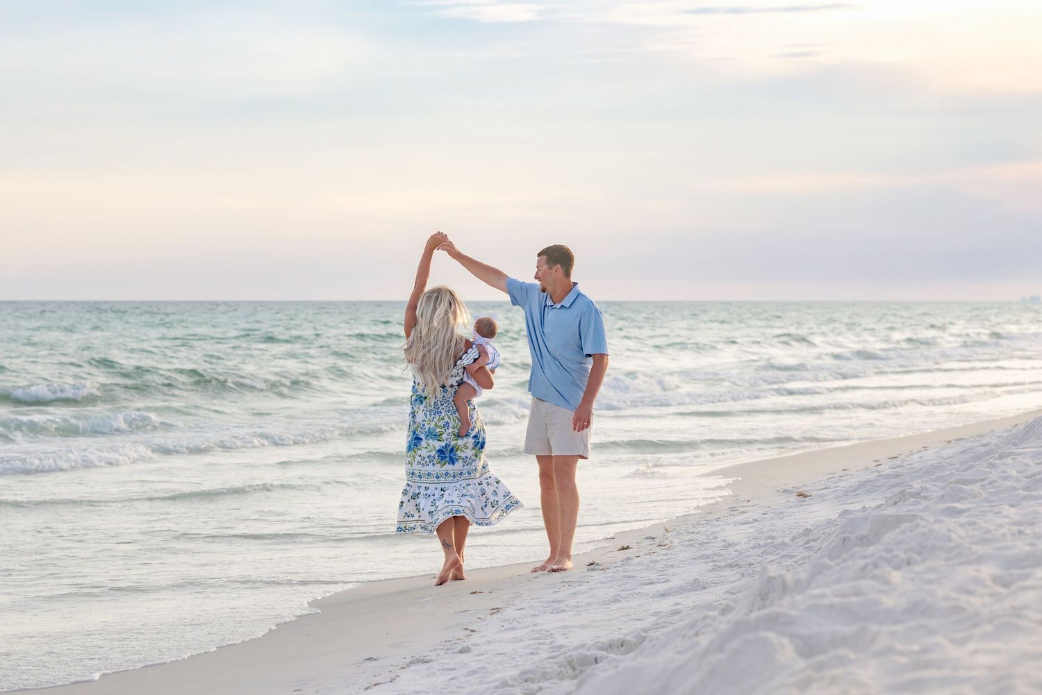 A couple dances on a sandy beach at sunset while holding a small child, with gentle ocean waves in the background.