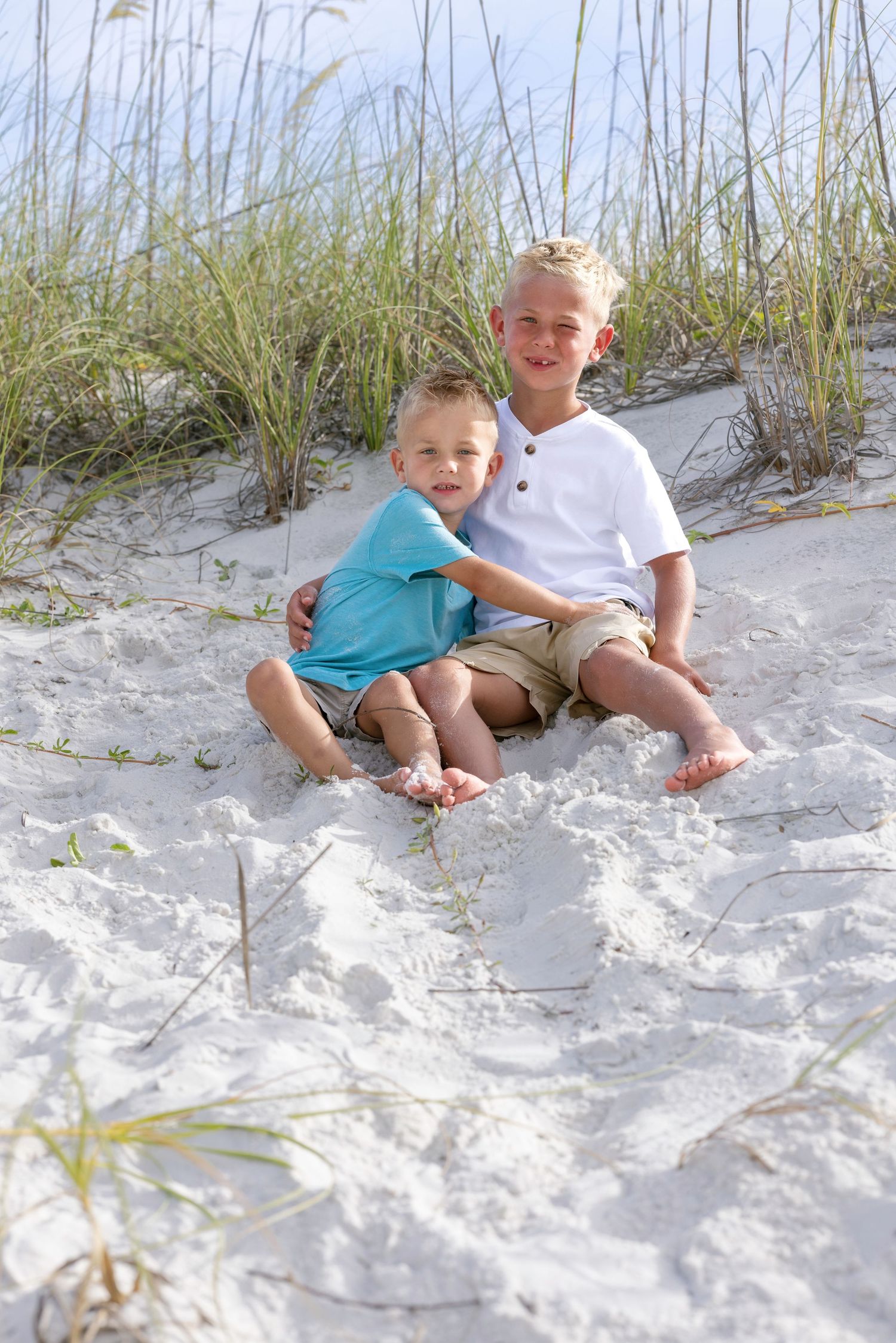 Two young boys sitting together on white sand dunes at the beach, smiling with sea grass in the background.