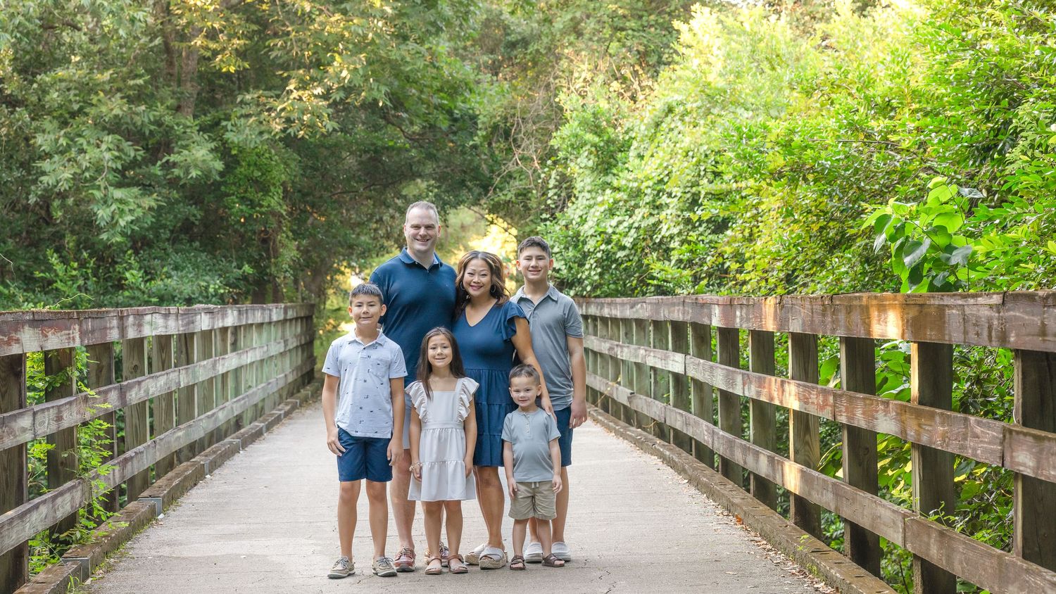 A family group walks together on a wooden bridge surrounded by lush green trees and foliage in a park setting.