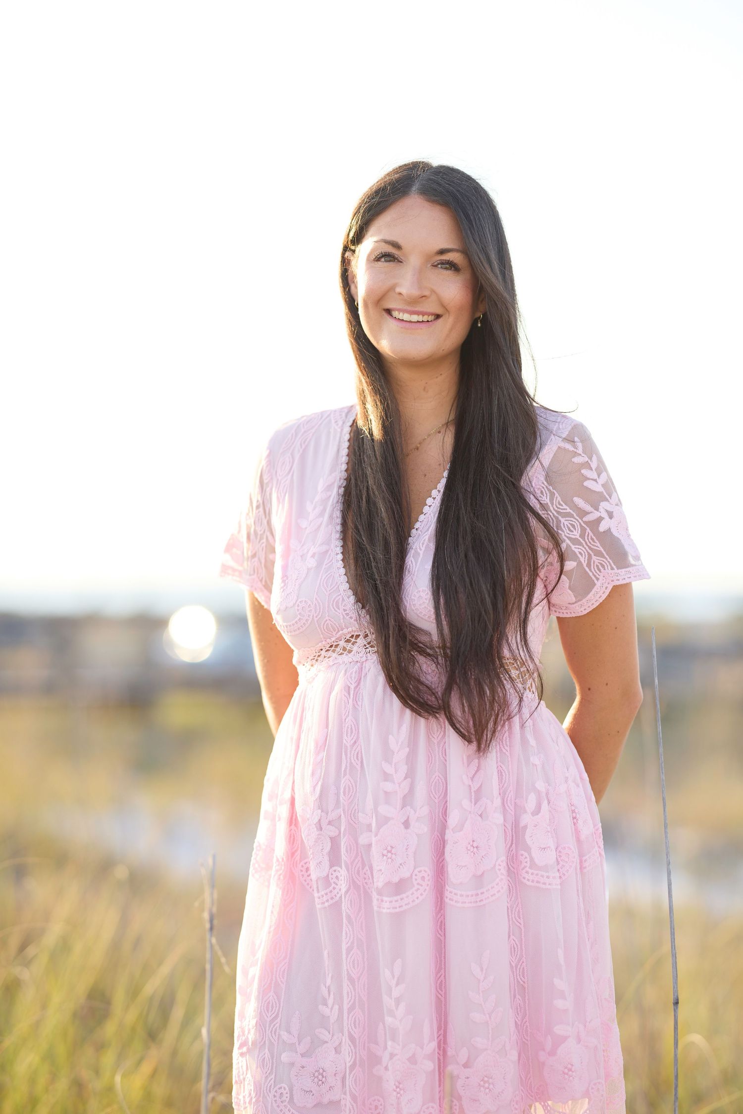 Smiling brunette woman in pink lace dress standing in golden field with bright sky background.