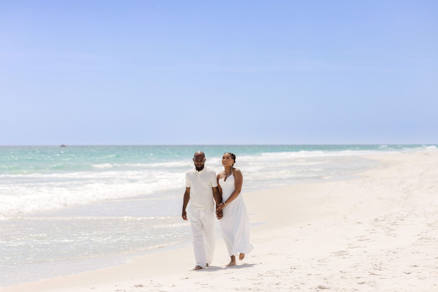 Couple in white outfits walking hand in hand on a sunny tropical beach with turquoise ocean waves.