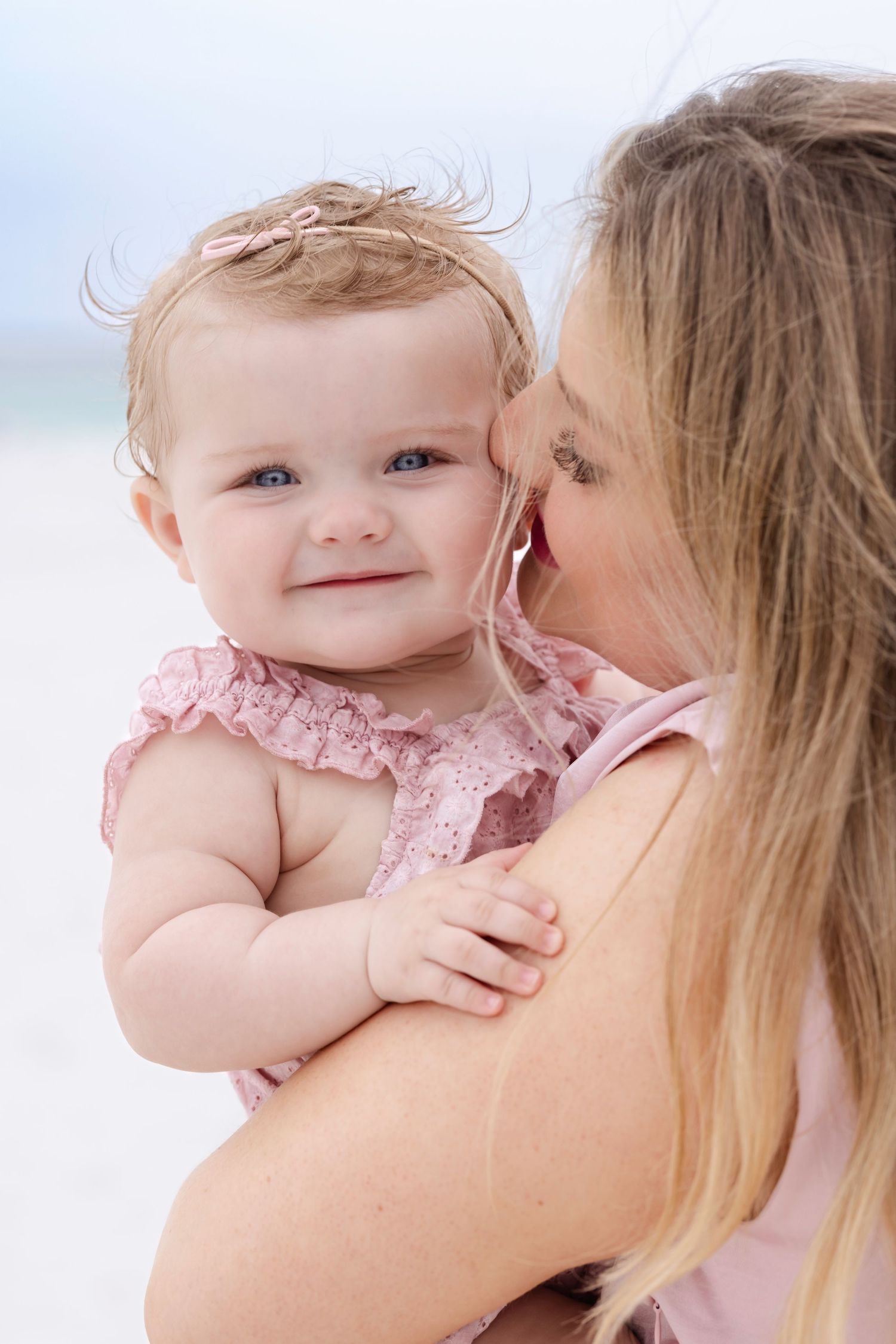 A mother kisses her smiling blue-eyed baby girl in a pink dress on a sunny beach.