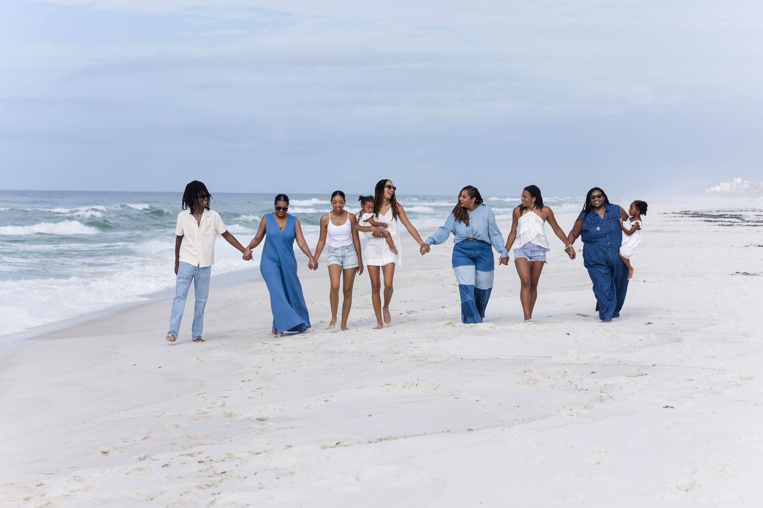Seven women holding hands walk along a sunny white sand beach with ocean waves in the background.