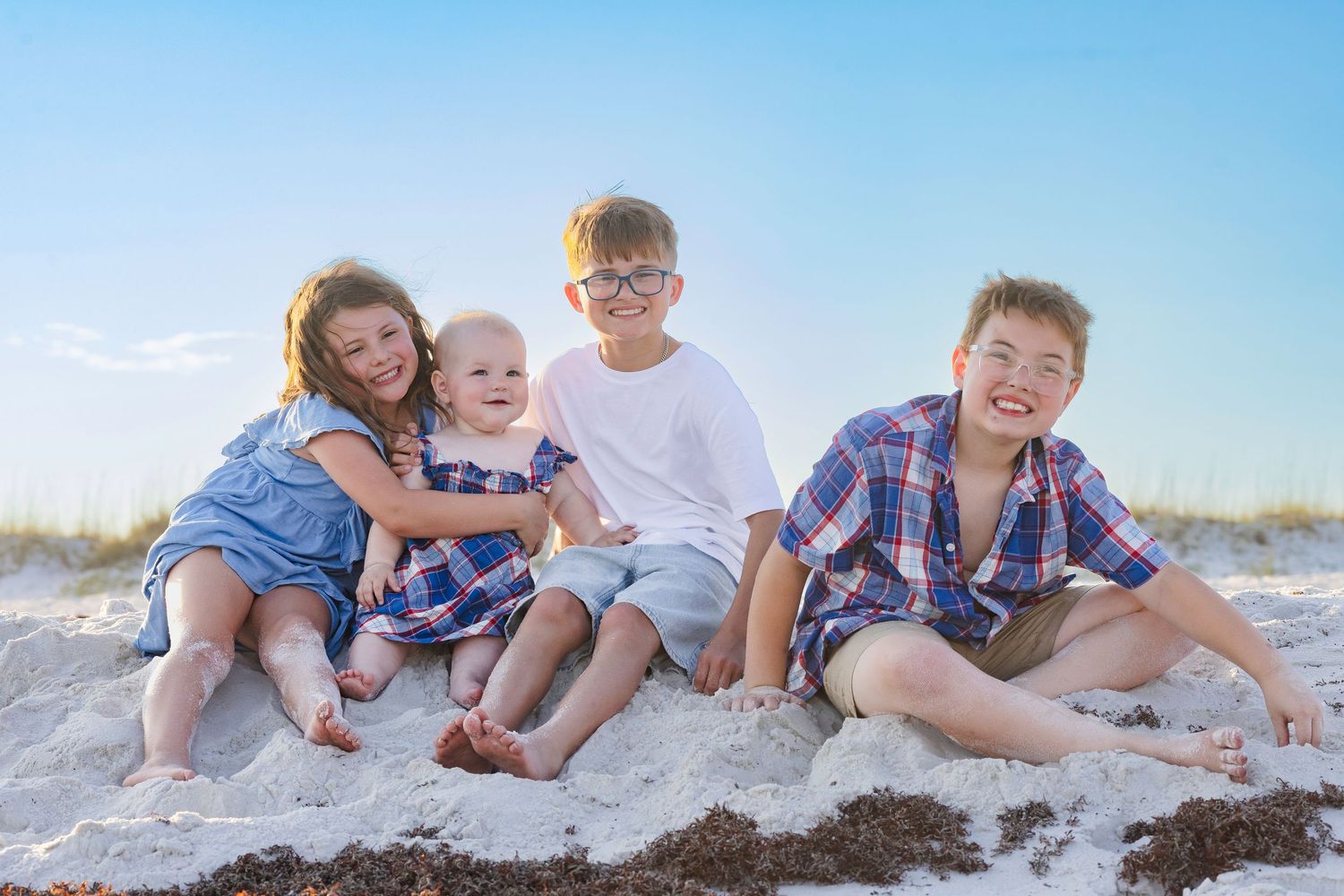 Four happy children sitting together on a sandy beach on a sunny day, smiling and posing for a family photo.