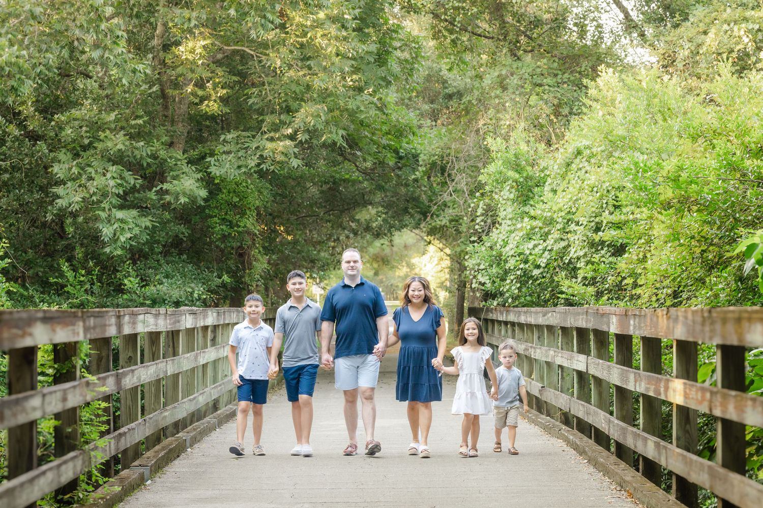 Family of six walking together on a wooden bridge surrounded by lush green trees in a park setting.