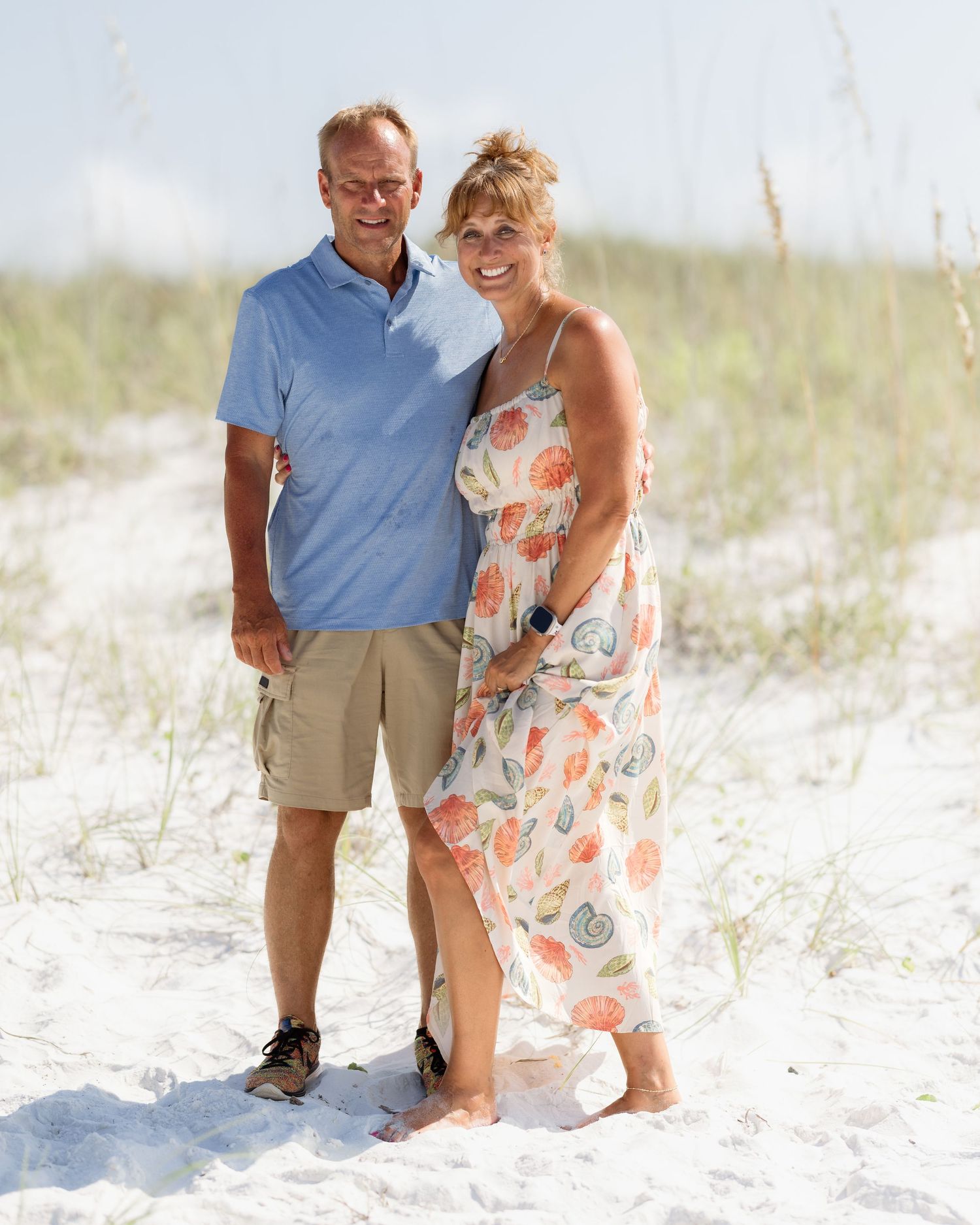 Smiling middle-aged couple standing together on sandy beach with sea oats behind them.