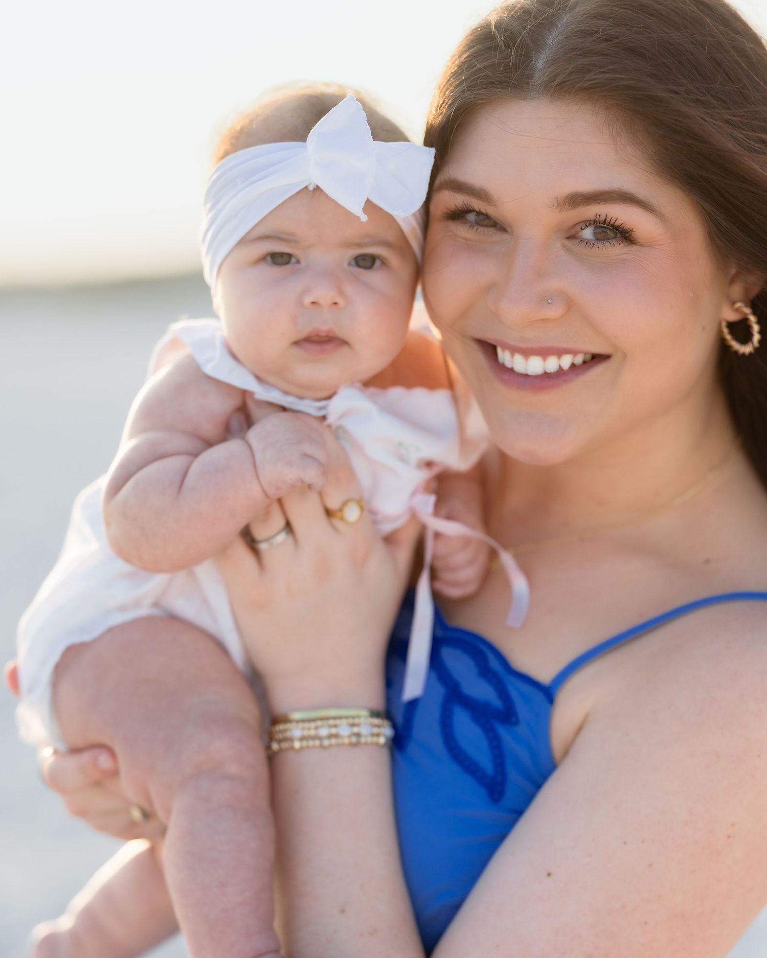 Smiling brunette mother in blue dress holding baby with white bow headband on beach.