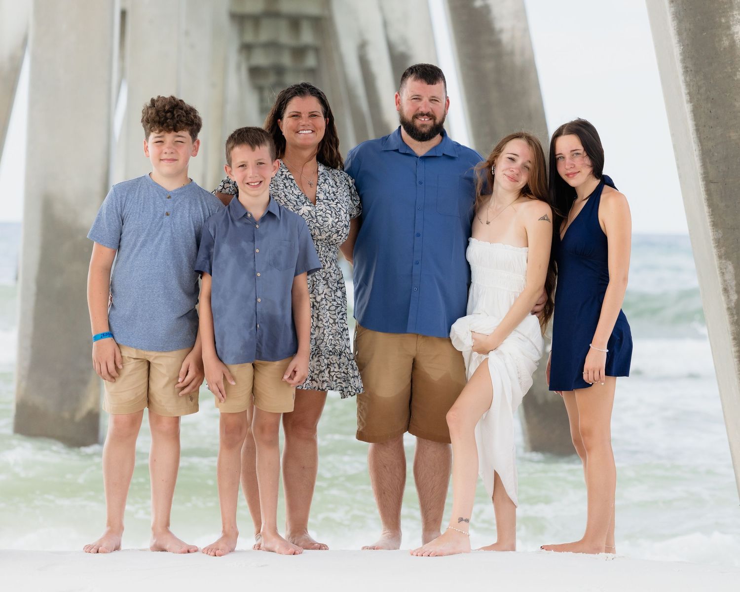 Family of six posing barefoot on beach near pier, parents with four children smiling in coordinated outfits.